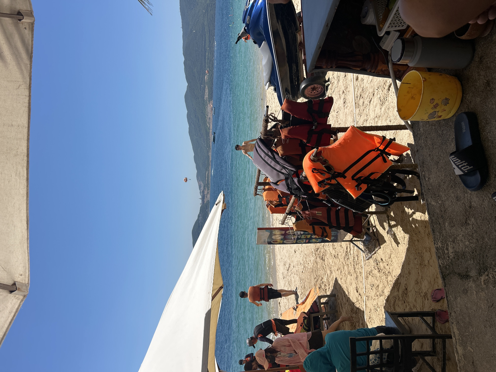 This image depicts a sunny day at a beach, likely in Nha Trang Ward, Vietnam, given the presence of a jet ski and the tropical scenery. The foreground shows a variety of life vests hanging on a rack, suggesting a water sports rental or activity area. A blue jet ski is parked on a trailer behind the life vests. On the beach, several people are visible, some wearing life vests, indicating they might be preparing for or have just finished water activities. One person stands in the shallow water, shirtless. In the background, the turquoise ocean stretches towards a mountainous coastline under a clear blue sky. A parasail is visible in the distance, pulled by a boat. The beach itself is sandy, with some lounge chairs and umbrellas providing shade. A white umbrella is prominently positioned in the upper left of the frame. The overall scene conveys a lively atmosphere of a tourist destination offering water recreation.