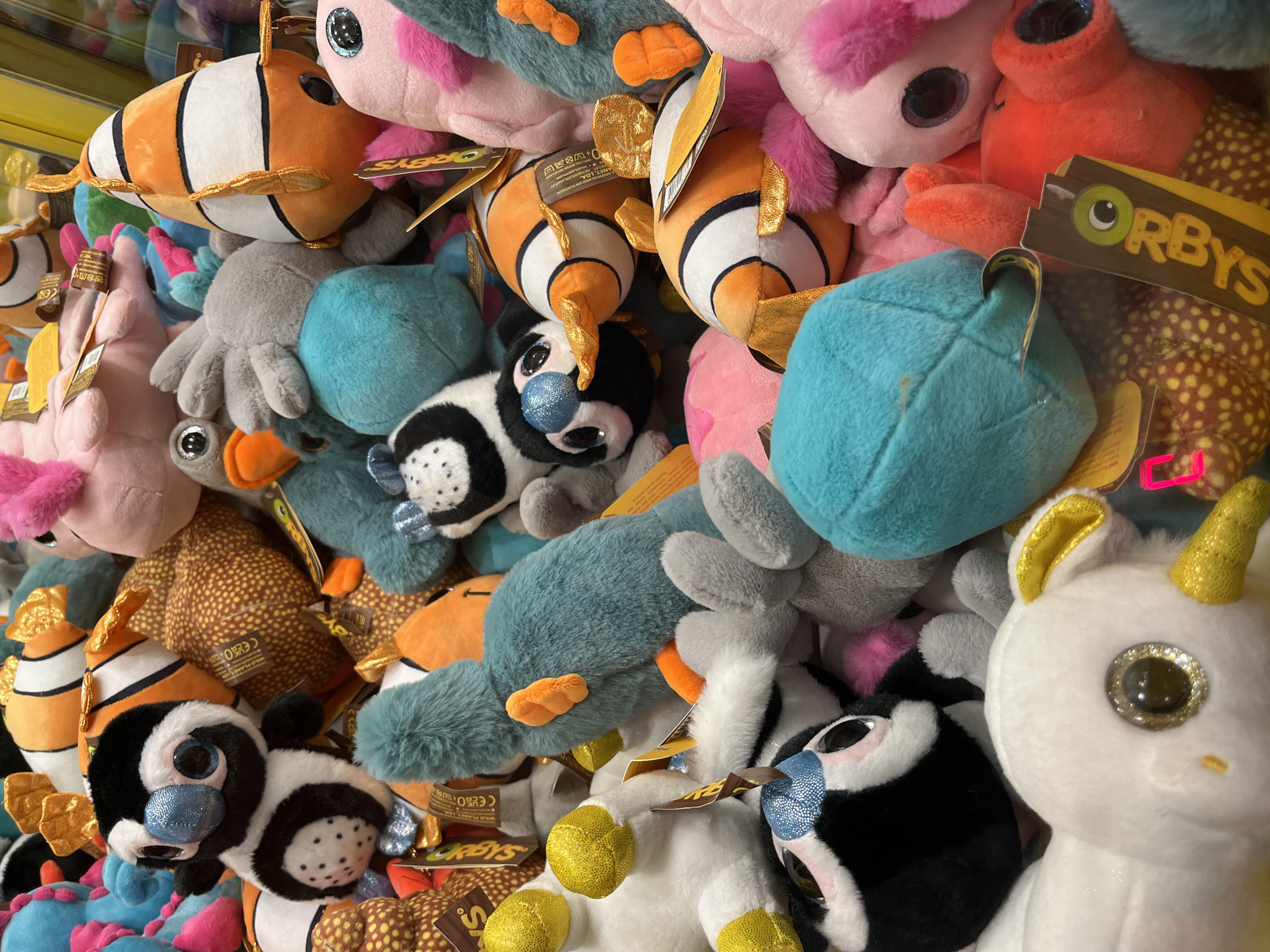 The image shows a close-up, overhead view of a collection of stuffed animals, predominantly sea-themed plush toys, within a glass display case. The toys include several clownfish, a penguin, a teal-colored platypus-like creature, a pink sea creature, a grey octopus-like creature, and other assorted soft toys in various colors like orange, white, pink, and blue. Many of the toys have large, shiny eyes. Some of the toys have tags attached, including one with the logo "ORBYS". The lighting suggests an indoor environment, likely a game arcade or amusement area, with a bright, reflective surface visible at the top. The stuffed animals are tightly packed together, obscuring much of the background. There are no people visible in the image, and no specific location in Budva, Montenegro, can be identified from the provided visual information. The scene is static, with the stuffed animals posed as they would be in a claw machine or display.