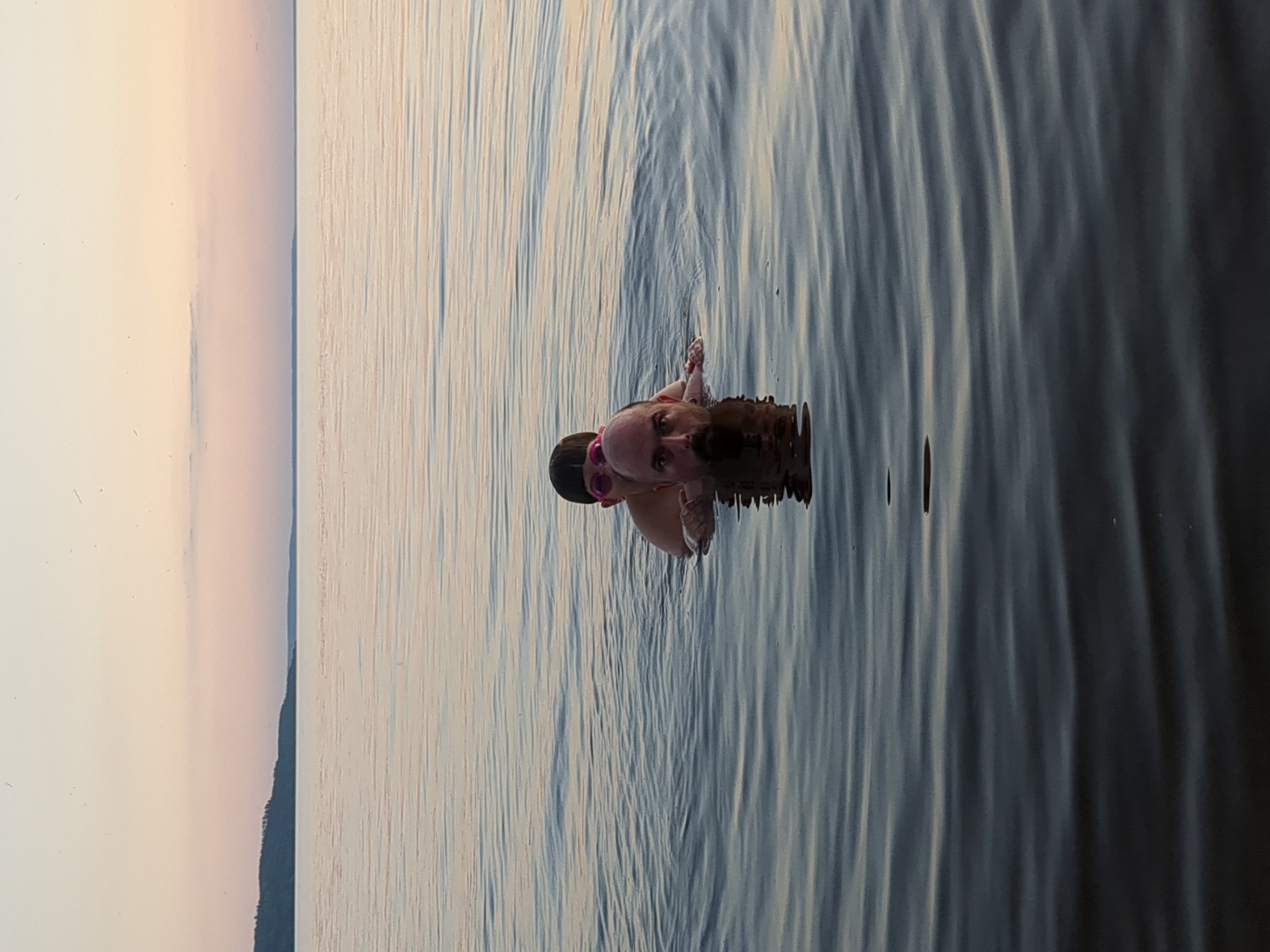 Two people are swimming in the water, a child with pink goggles on top of an adult. The adult's head and shoulders are visible above the water, as is the child's head. The background shows a horizon with a faint landmass, suggesting a large body of water such as a lake or sea. The sky is clear with pastel colors indicating dawn or dusk. The water has gentle ripples. The image was taken at the Kyiv Reservoir, part of the Dnipro River, near the city of Pereiaslav, Ukraine.