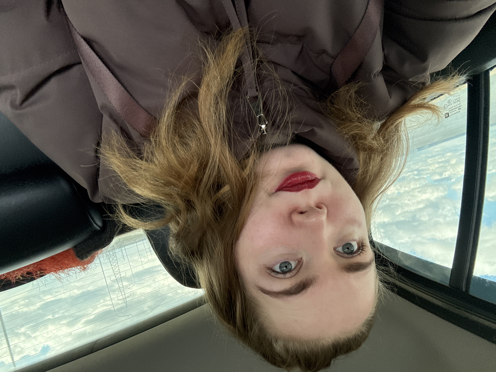 A young woman is captured in a selfie taken inside a car, looking directly at the camera with a neutral expression. She has light brown to auburn hair and striking blue eyes, accentuated by red lipstick. She is wearing a dark brown, puffer-style jacket, and a safety belt strap crosses her chest.
The setting is inside a vehicle, likely in the back seat, as indicated by the headrest visible on the left and the rear window providing the backdrop. Through the car's rear window, a bright, cloudy sky with scattered white clouds is visible, suggesting daytime. Faintly visible beyond the clouds, and possibly reflected in the window, are what appear to be power lines or a distant structure. On the car window, several markings are visible, including "SBRTOO O 123", "DOT", "AS2", "M129", and partially "TRANSPARENT," which are typical automotive glass certifications. An orange, fluffy object, possibly a blanket or pillow, is also visible near the bottom left of the window frame.
The overall scene suggests a moment of rest or travel, with the subject taking a picture from within the car. The specific location context is Noviy, Russia.