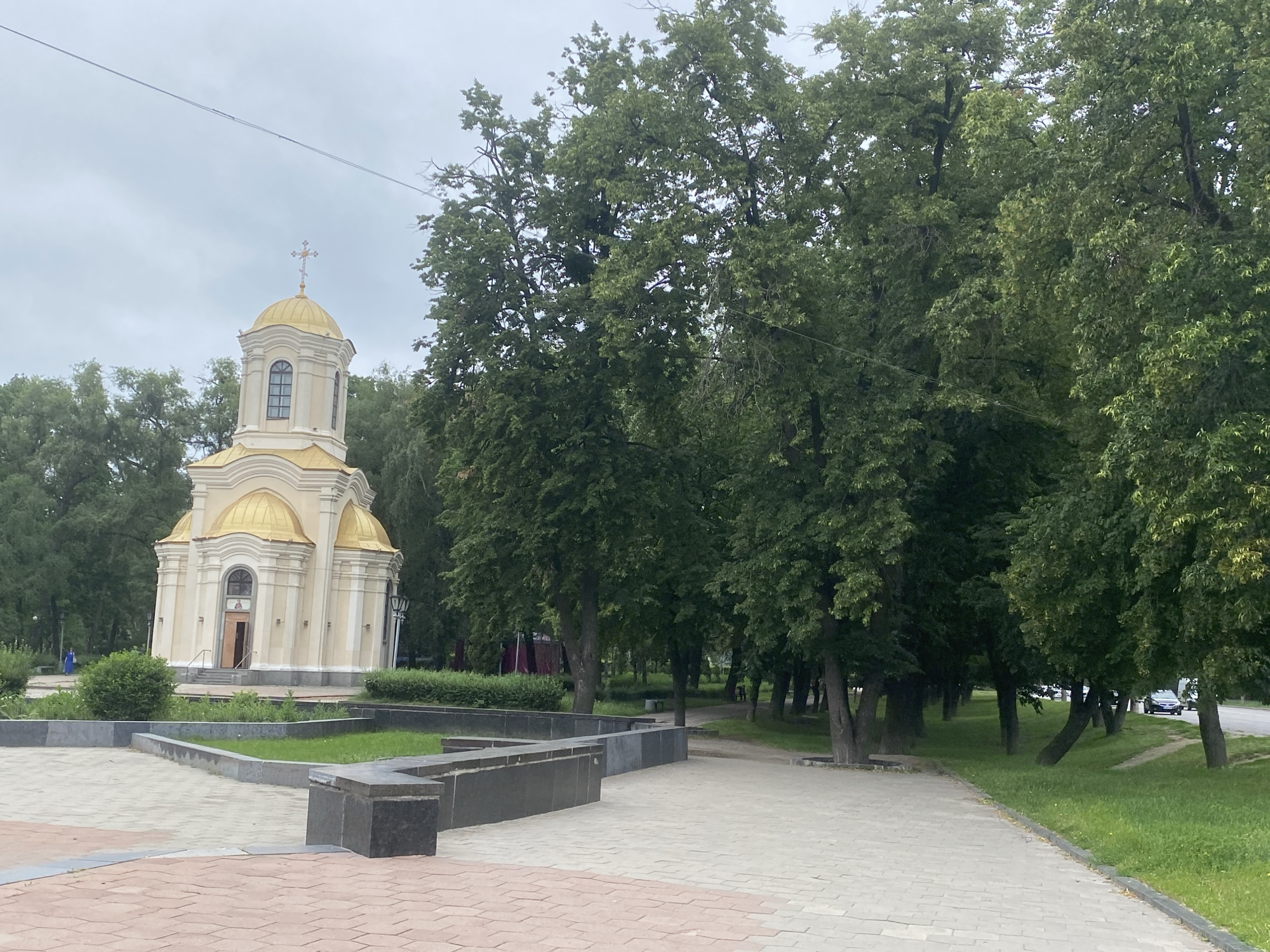 The image depicts a small, cream-colored church with a golden dome and a cross on top, located in Poltava, Ukraine. The church has arched windows and a simple, classical architectural style. It is surrounded by lush green trees and a paved walkway. In the foreground, there is a gray stone planter with grass and hedges. A few people are visible in the distance, and a car is parked on a road behind the trees. The sky is overcast, suggesting a cloudy day. The scene appears to be a public park or square adjacent to the church.