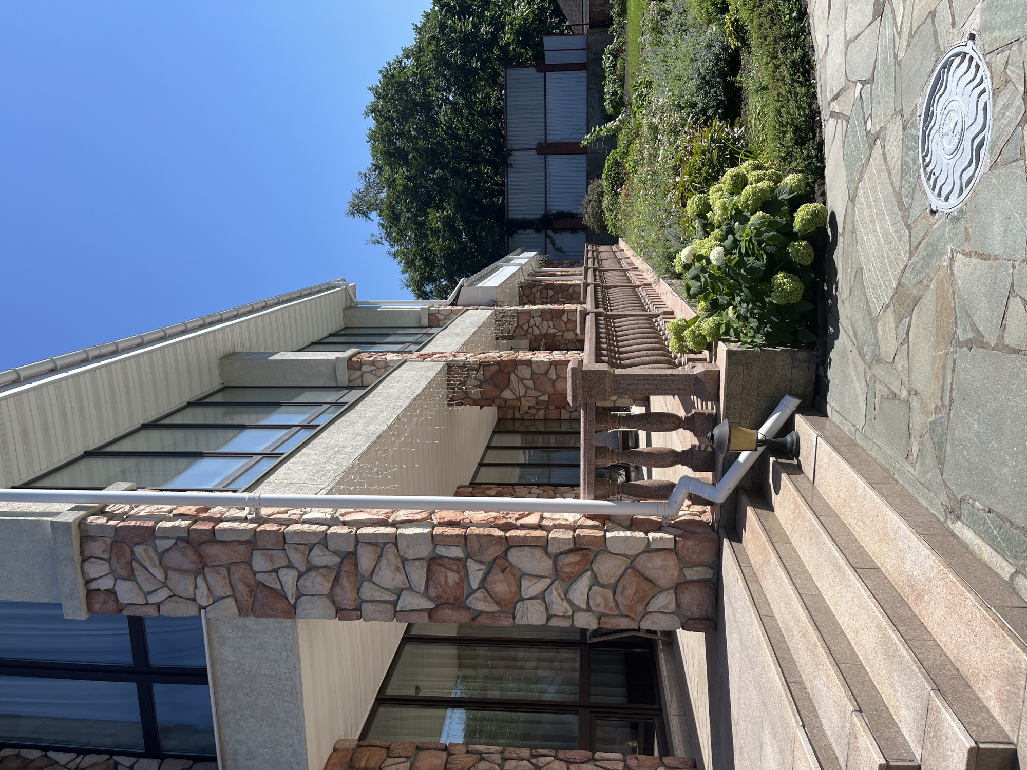 The image shows a view of a modern house with stone-clad columns and a porch, located in Vladivostok, Russia. The house has large windows and a white soffit. A downspout runs along one of the stone columns, directing water towards a small lamp fixture on the steps. The steps themselves are made of light-colored stone with a flecked pattern. In the foreground, a pathway is laid with irregular-shaped gray flagstones, and a round metal manhole cover with a wavy pattern is visible. To the right of the steps and pathway, a landscaped garden area features lush green bushes, including a large hydrangea bush with white blooms, and various other plants. A metal fence is visible in the background, behind the garden. The sky is clear and blue. No people are visible in the image.
