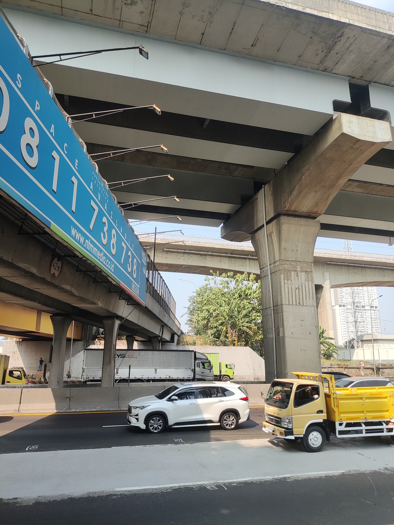 The image depicts an overpass in Lambangsangi, Indonesia, with a blue billboard advertising available space and contact information. Beneath the overpass, a white SUV and a yellow flatbed truck are traveling on a highway. In the background, other vehicles, including a white semi-truck, are visible. The scene is characterized by concrete structures, traffic, and lush green trees.