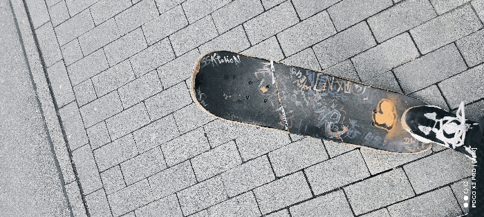 The image displays a high-angle view looking down at a single person's left foot positioned on a skateboard, set against an outdoor paved surface. The skateboard, dark in color, exhibits significant wear, with numerous light-colored scuffs, graffiti-like markings, and a prominent yellow-orange irregular blotch on its griptape surface. The tail and nose areas show visible chipping and abrasion. Four circular holes are present near the center of the board.
The foot is clad in a black athletic shoe with white laces and white accents, its sole firmly pressed onto the skateboard.
The ground consists of grey rectangular paving stones laid in an interlocking pattern. To the upper left of the frame, there is a distinct transition line separating the paving stones from a darker, fine-grained, asphalt-like surface. No other individuals or specific landmarks are visible. The scene depicts an urban outdoor environment, but the precise location within the city of Constance, Germany, is not determinable from the provided visual information.