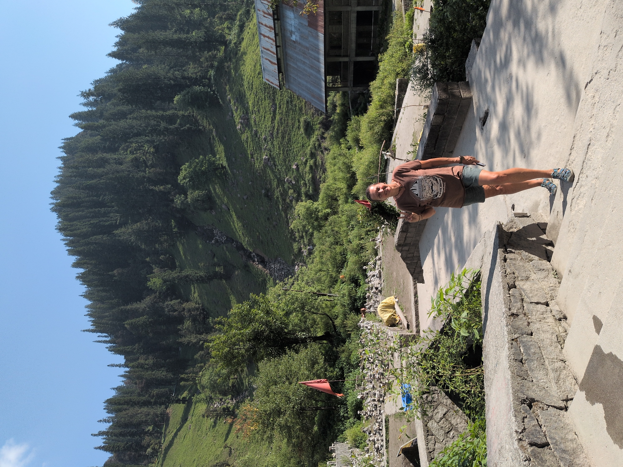 A woman stands on a concrete path in Majhach, India, giving a peace sign to the camera. She is wearing a brown t-shirt with a graphic print, green shorts, and blue sandals. Behind her, a lush green mountainside is covered in dense pine trees. A rustic, weathered building with a tin roof is visible on the right side of the frame. In the foreground, stone walls and steps lead down to a path, and a small orange flag is planted in the ground among a collection of stacked stones. The sky is a clear blue.