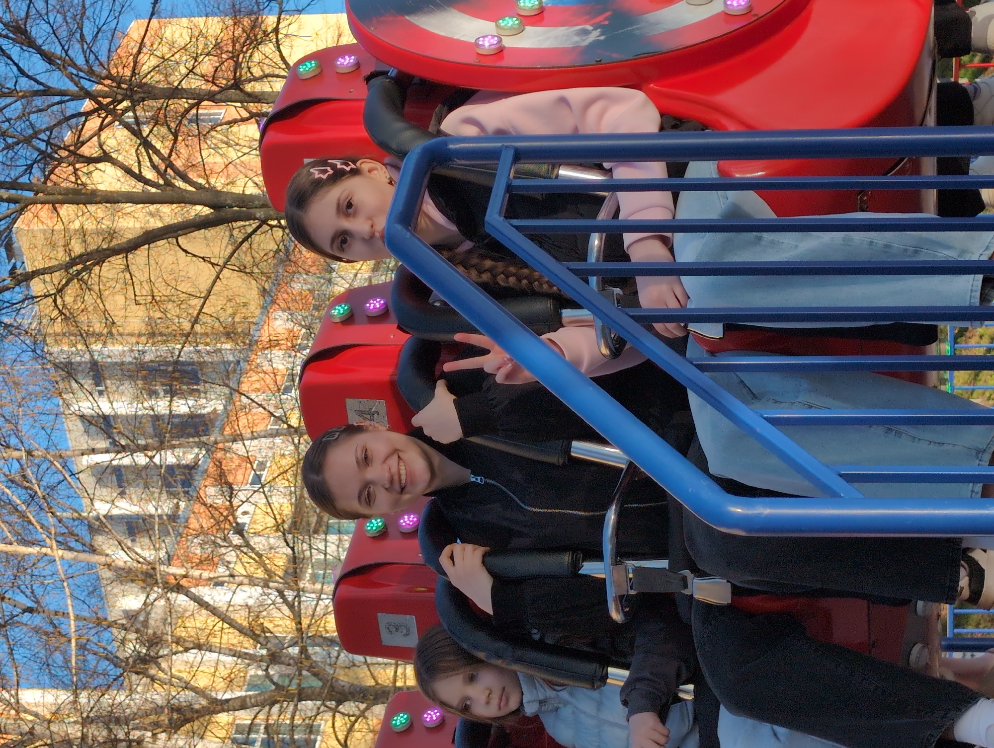 Three girls are seated on a spinning amusement ride, with a red base and blue safety bars. The ride has colorful lights on its sides. The girl in the middle is smiling and holding up a peace sign. The background includes trees and a building with a yellowish facade. The photo was taken in Izhevsk, Russia.
