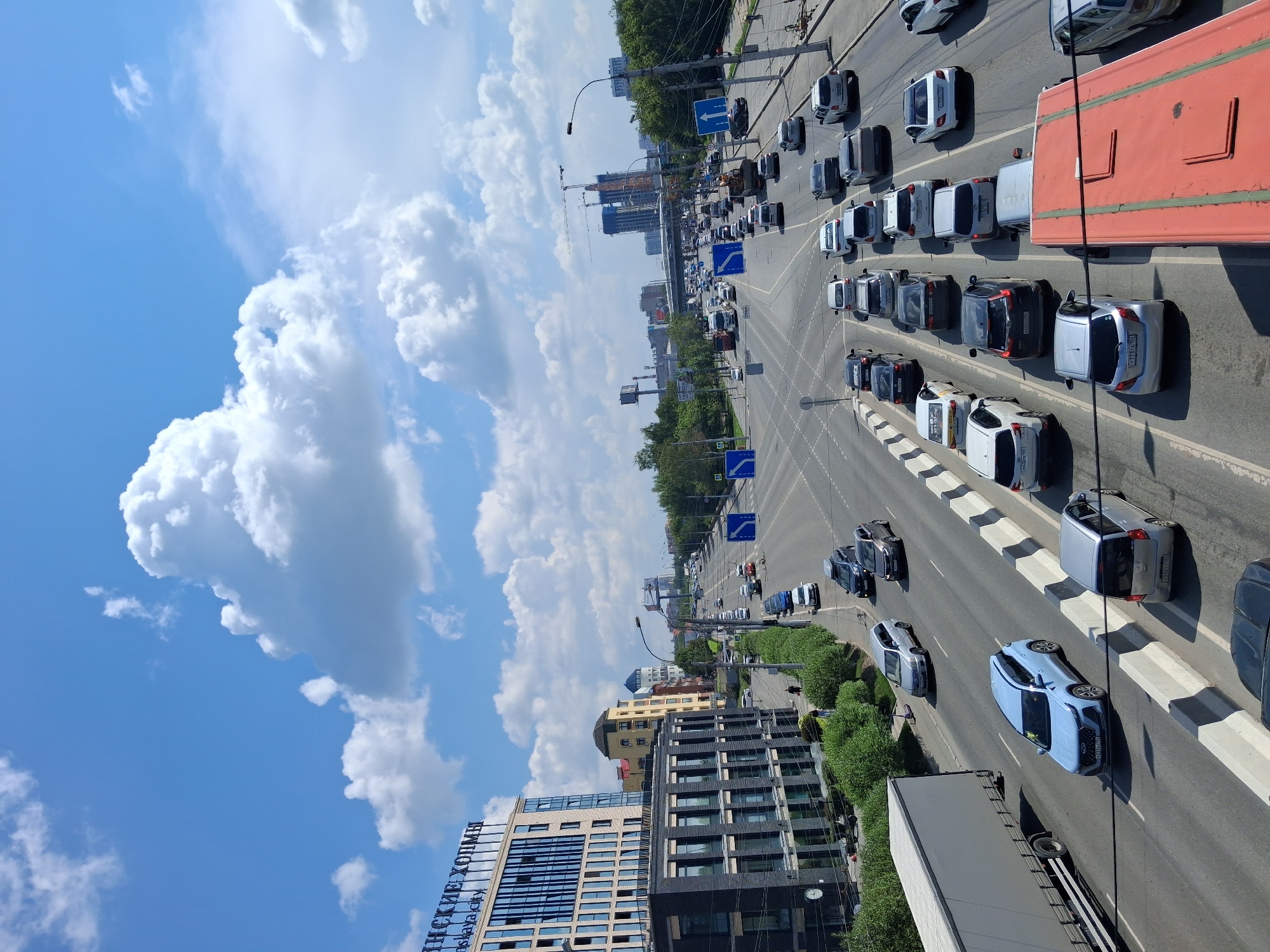 This is a medium-angle, slightly elevated, exterior, eye-level, full-shot, horizontal, color photograph of a busy city street in Novosibirsk, Russia, on a sunny day with scattered white clouds. The scene is dominated by traffic on a multi-lane highway. A light blue car is in the foreground, on the left side of the road, heading towards the viewer. To its left is a large, grey semi-trailer truck. Further down the road, a long line of cars is visible, moving away from the viewer. The road is divided by white lines and concrete barriers. On the left side of the road, there is a modern, multi-story building with glass windows and signage that reads "ИНСКИЕ ХОЛМЫ". Next to it is a darker, modern building. On the right side of the road, trees and more buildings are visible in the distance, including some under construction with cranes. Several blue traffic signs are posted along the road, indicating traffic flow. There are no people clearly visible in the foreground, but a few figures can be seen walking on the sidewalk on the left side of the road. The sky is a clear blue with fluffy white clouds.