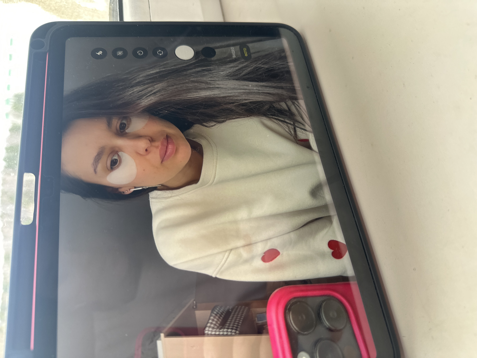 A photograph shows a cell phone displaying an image of a woman with dark hair, wearing white under-eye patches, a light-colored sweater with red heart decals, and an earbud in her left ear. The phone is on a surface with a white-painted wall behind it. A reflection in the phone screen reveals a drawer with items in it, including a checkered object and a pink phone case. The location is unknown and cannot be determined to be Chaikovsky, Russia.