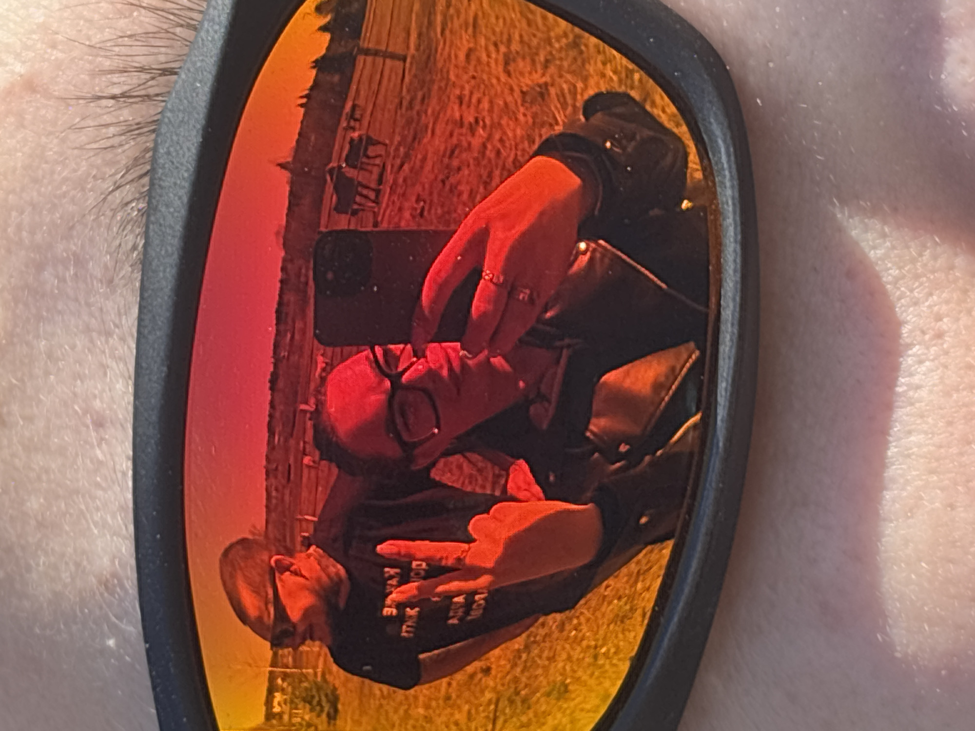 The image shows a close-up of sunglasses reflecting a person taking a selfie. The person is wearing glasses, a black t-shirt with white text, and a leather jacket. They are holding a smartphone with one hand, fingers in a "peace" sign, and using the other hand to take a photo. The background reflected in the sunglasses appears to be a grassy field with a wooden fence. The location is likely rural, but it is not possible to confirm if it is in Opary, Russia. The edges of the sunglasses frame and a portion of the photographer's eyebrow and cheek are also visible.