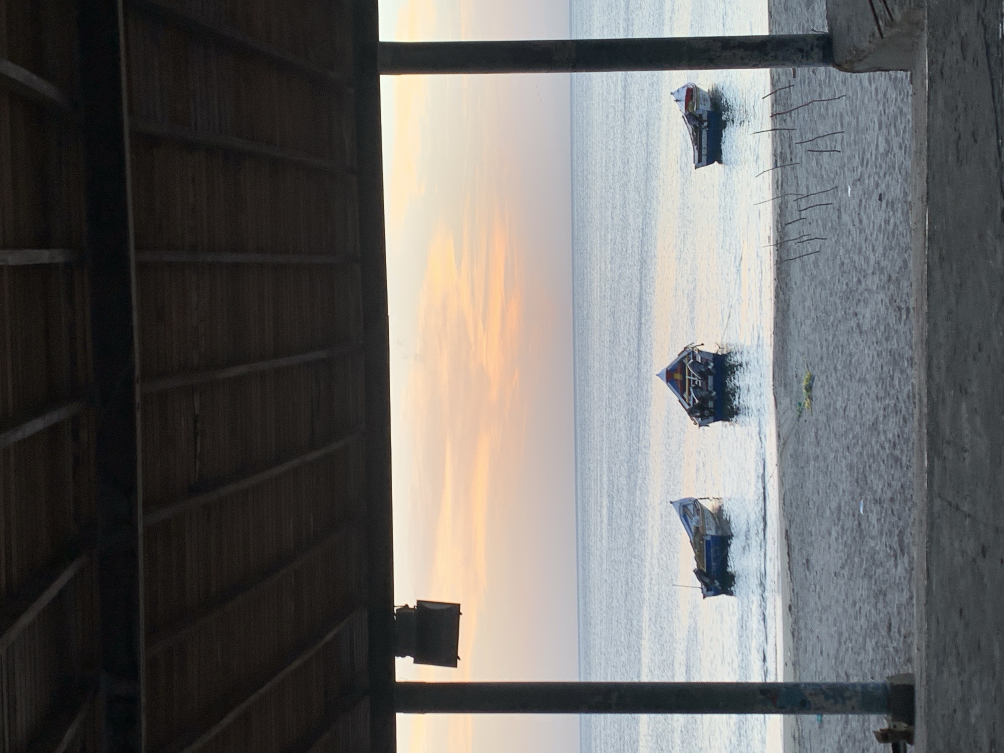 The image, taken in Juan Griego, Venezuela, depicts a serene coastal scene at dusk. Framed by a rustic wooden canopy and concrete structures, the view opens to a calm sea under a pastel sky painted with hues of orange and pink. Three small fishing boats are moored in the shallow water near the sandy shore, appearing idle. The beach itself is sparsely populated, with only a few scattered pieces of debris and some upright sticks creating a subtle pattern on the sand. The overall atmosphere is one of tranquility and quietude, typical of a coastal town at the end of the day.