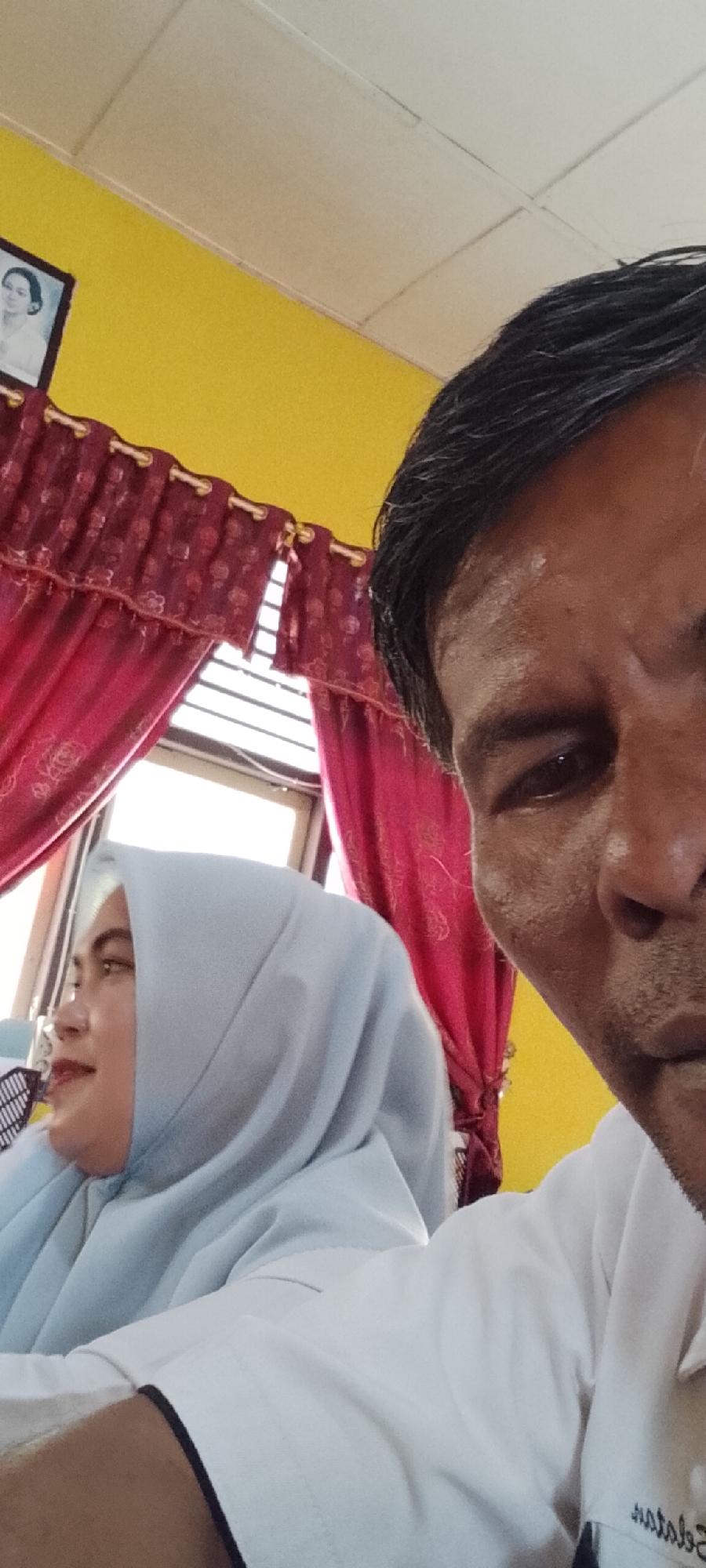 In a room with yellow walls and a white tiled ceiling, a man and a woman are visible. The man is in the foreground, taking a selfie, with his right eye, nose, and forehead in focus. He has dark, short hair and appears to be sweating. He is wearing a white collared shirt.
To his left and slightly behind him, a woman is visible from the shoulders up. She is wearing a light blue hijab and looking to her left. Her skin tone is lighter than the man's.
Behind them, red curtains with floral patterns hang over a window with blinds. On the wall above the curtains, a framed black and white photograph of a woman in traditional attire is displayed. The overall impression is of an indoor setting, likely a home or office in Jepara, Indonesia, given the context provided.