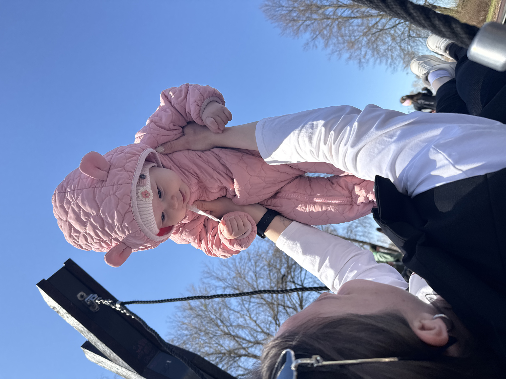 A low-angle shot depicts a baby in a pink snowsuit with bear ears being held aloft against a clear blue sky. The baby wears a white hat with a flower and is looking downward with a slight smile. Below the baby is a person wearing a white long-sleeved shirt and a black watch on their left wrist. The person's head is partially visible with dark hair and sunglasses perched on top of their head. A portion of a black object with metal attachments and chains is visible on the left. In the background, bare trees are visible, along with other individuals wearing white shoes and black pants. The location is Zav'yalovo, Russia.