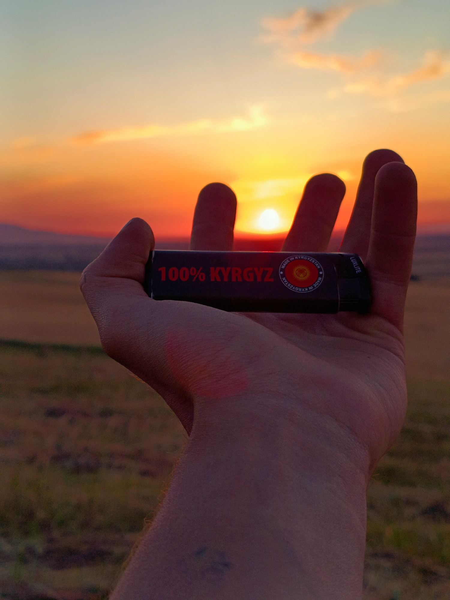 A close-up, shallow depth-of-field shot captures a person's open palm holding a dark object against a sunset. The object, a lighter with red text "100% KYYGZ" and a circular emblem, is positioned horizontally. The sunset fills the background with warm hues of orange and yellow, with a bright sun visible in the distance. A blurred landscape of rolling hills and dry grass stretches beyond the hand. The setting appears to be a rural outdoor location, potentially in Arashan, Kyrgyzstan, given the text on the object.