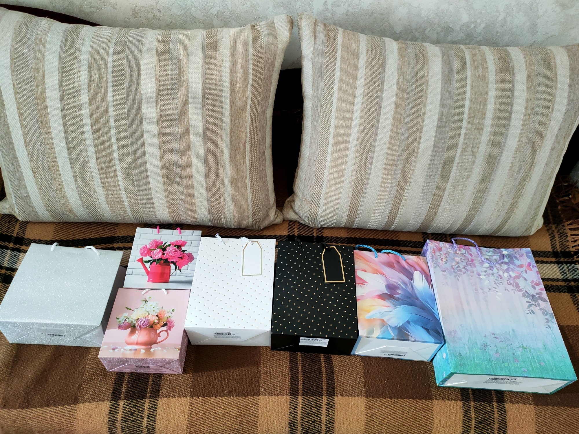 This media file displays a collection of gift bags arranged on a patterned blanket, which is likely a couch or bed. Two large pillows with vertical beige and white stripes are visible in the background, resting against a textured wall.
The subjects are the gift bags themselves, which are varied in design. From left to right, there is a large, plain silver bag with a glittery finish. Next to it is a smaller, square pink bag with an illustration of a bouquet of flowers in a pink teapot. To the right of that is a white bag adorned with small gold polka dots and a gold tag. Following that is a black bag with the same gold polka dot pattern and a black tag. The next bag features a vibrant, abstract floral or feather design in shades of pink, blue, and orange. The last bag, on the far right, is a tall, rectangular bag with a soft watercolor-style illustration of foliage and abstract floral elements in pastel blues, purples, and greens. All the bags have handles, and some have visible barcodes and text on their lower portions.
The scene appears to be indoors, likely in a living room or bedroom, given the presence of the pillows and blanket. There is no discernible activity taking place; the gift bags are simply displayed. The lighting suggests it could be daytime, but no specific time of day can be determined. There are no people visible in the image.
Notable details include the diversity in the gift bag designs, suggesting a variety of occasions or recipients. The overall impression is one of preparation for gifting or a celebration. No emotions are explicitly conveyed. The visible text on the gift bags is primarily informational, such as barcodes and possibly manufacturer details, and is not clearly legible for specific interpretation.