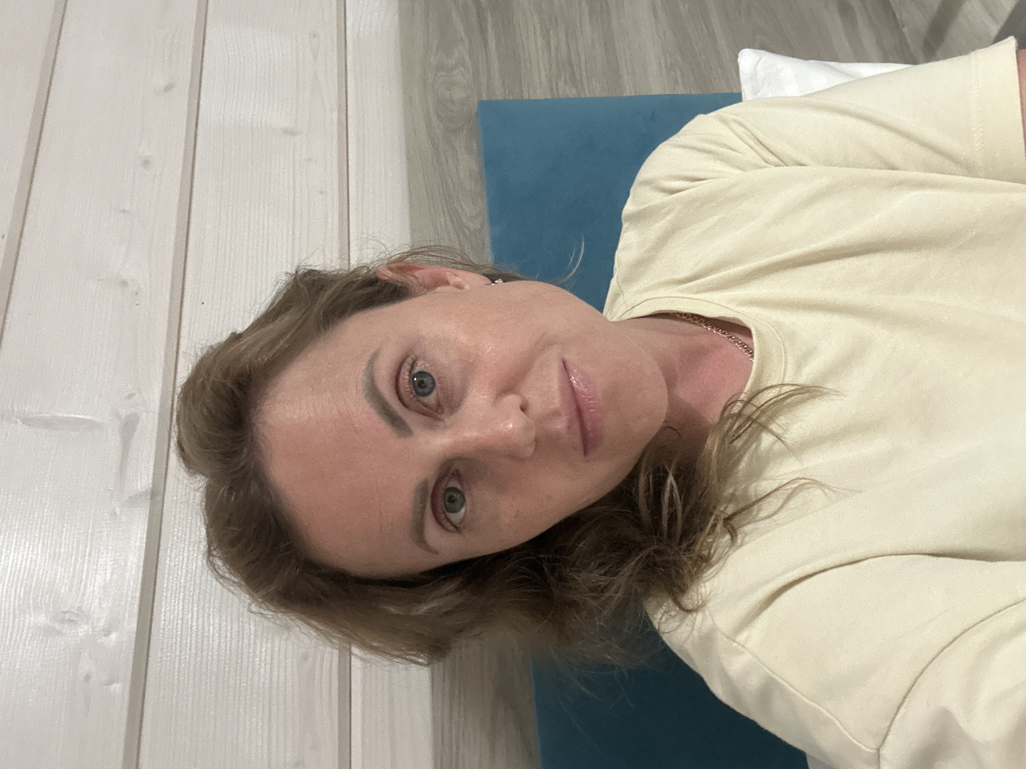 The image shows a medium shot of a woman with medium-length brown hair, looking directly at the camera. She is wearing a light yellow t-shirt and a necklace. She is positioned in front of a wall covered with white horizontal wooden planks, with a teal-colored headboard behind her. A white pillow is partially visible to her right. The scene is indoors and appears to be a bedroom. There are no discernible landmarks or features that would specifically place this image in the city of Anapa, Russia.