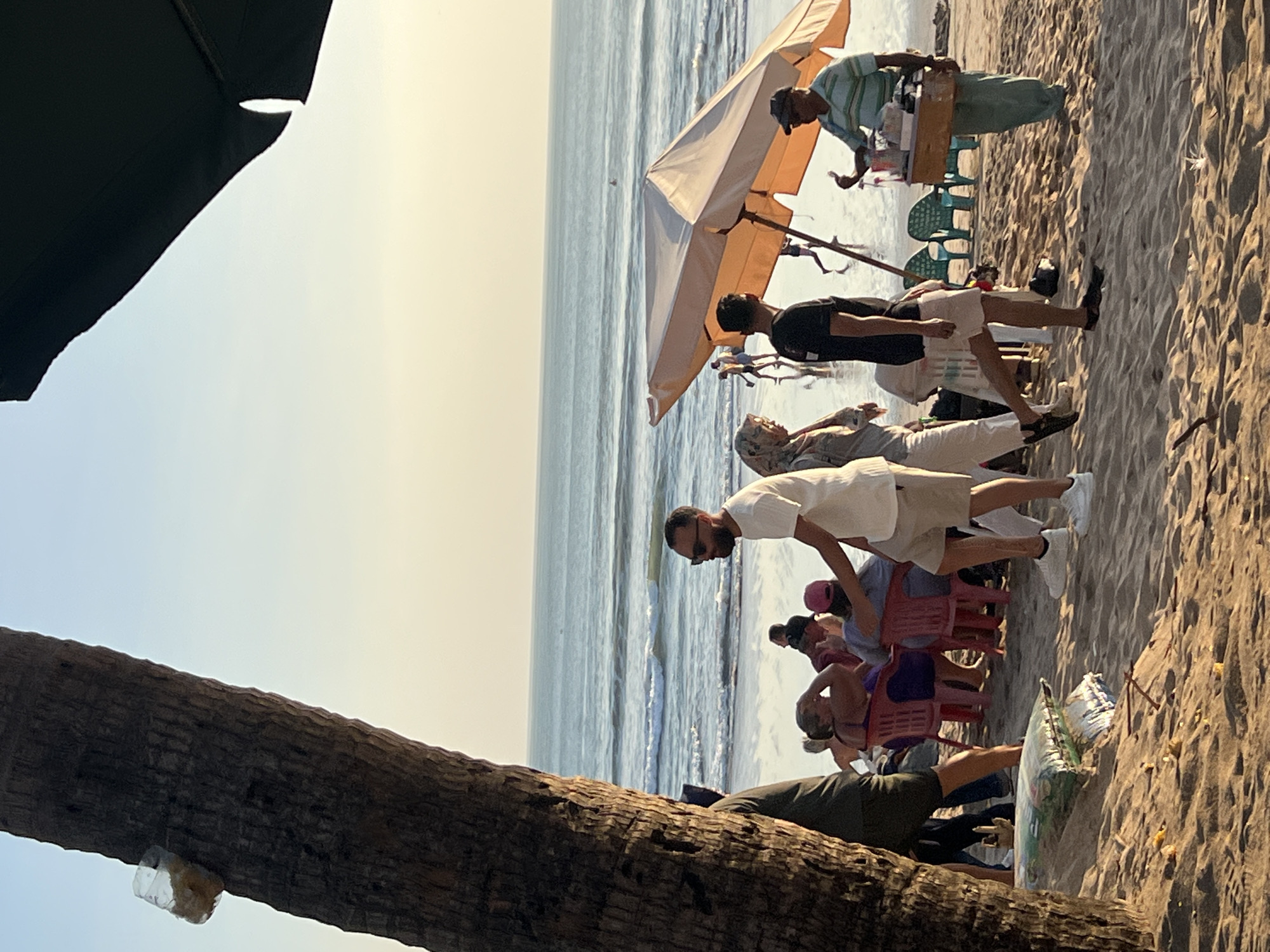 The image depicts a lively beach scene in Kuta, Indonesia, during the late afternoon. The foreground is dominated by a large palm tree trunk on the left, with a clear plastic bottle containing some yellow material attached to it. The sand is a prominent element, with several people gathered in small groups.
To the right of the palm tree, a man in a white t-shirt and khaki shorts stands facing away from the camera, looking towards the ocean. In front of him, a group of people are seated on red chairs. A large, light-orange beach umbrella is set up, casting a shadow over some of the people and a vendor.
The vendor, a man in a striped polo shirt and green pants, stands behind a wooden cart filled with drinks and possibly food items. He is interacting with another person, possibly a customer, with his hand raised.
In the middle ground, the ocean stretches out to the horizon, with gentle waves breaking on the shore. Several individuals can be seen walking and playing in the shallow water. The sky is a soft gradient of light blue and pale yellow, indicating the approaching sunset. In the background, the silhouette of a distant surfer can be seen on a wave.
Scattered on the sand are a few items, including a large bag that appears to be some sort of packaging or merchandise. The overall atmosphere suggests a relaxed and social beach environment.