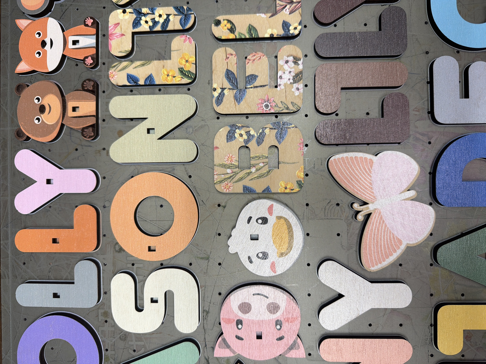 The image shows a close-up view of a wooden alphabet puzzle board. The puzzle pieces are cut out in the shapes of letters and some animal characters. The letters are multicolored and some are decorated with floral patterns. The animal characters include a bear, a fox, a pig, a duck, and a butterfly. The puzzle is set against a dark gray background with visible circular indentations and small holes, suggesting it is a pegboard or a similar display system. The lighting is even, highlighting the textures of the wood and the colors of the puzzle pieces. There are no people or any indications of a specific location in the city of Ternopil, Ukraine.