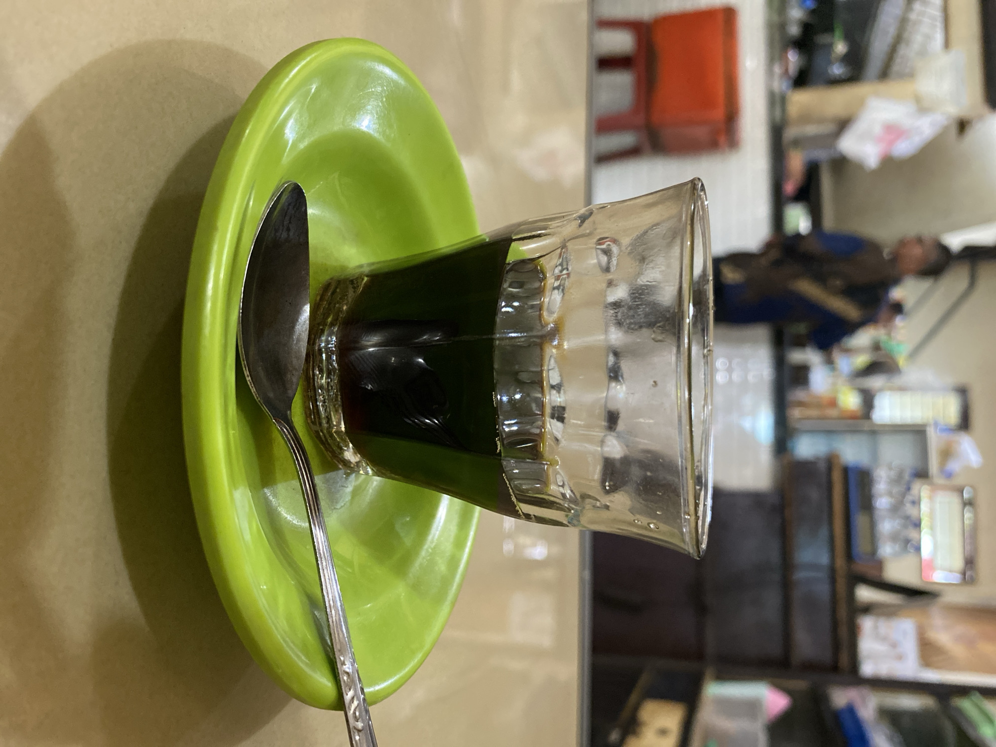 A clear glass of dark liquid, likely coffee, sits on a bright green saucer. A silver spoon rests across the saucer, partially under the glass. The table surface is light brown and appears slightly reflective. In the background, out of focus, is what appears to be a food stall or cafe. A person wearing a dark blue garment with a patterned sash is visible behind a counter. There are red containers and shelving with various items in the background. The scene is indoors, suggesting a casual dining or coffee shop environment. The presence of "Lhokseumawe, Indonesia" in the prompt strongly suggests the location.