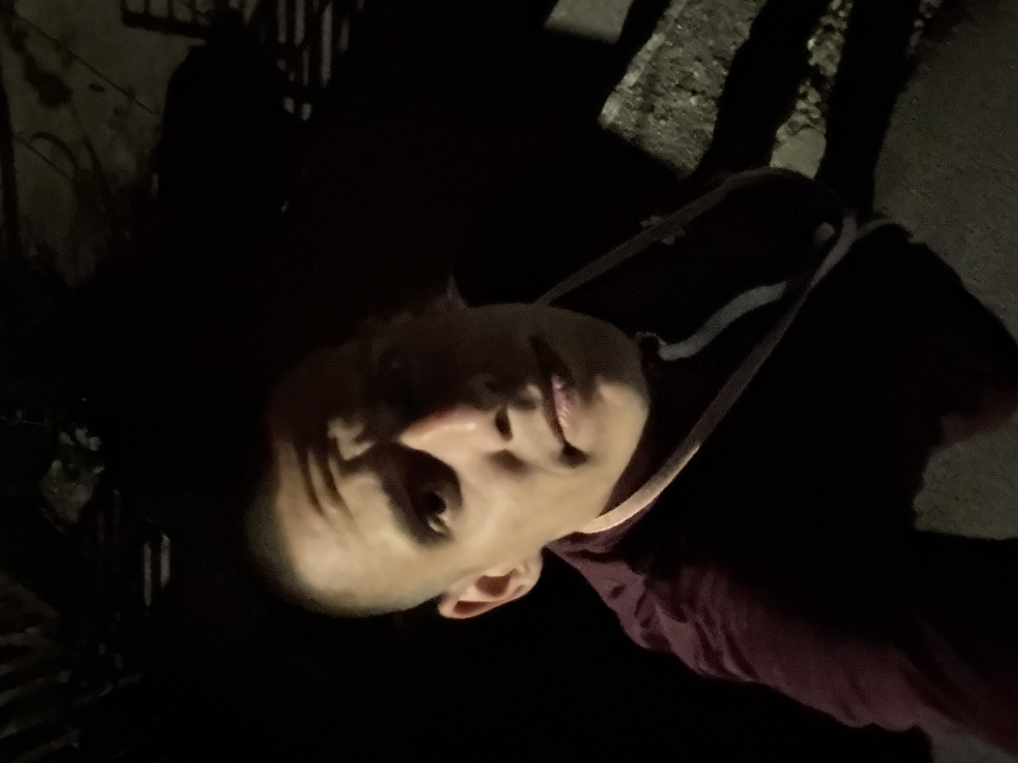 A headshot of a bald man, potentially of Caucasian descent, is seen with his head tilted to the left. The photograph appears to be taken at night with limited lighting. The man is wearing a dark garment with a white stripe across his chest, with a reddish-purple sweater underneath. The background includes what appear to be shadows of a fence and a concrete surface, potentially a sidewalk. No specific location in Sofia, Bulgaria can be determined with certainty.