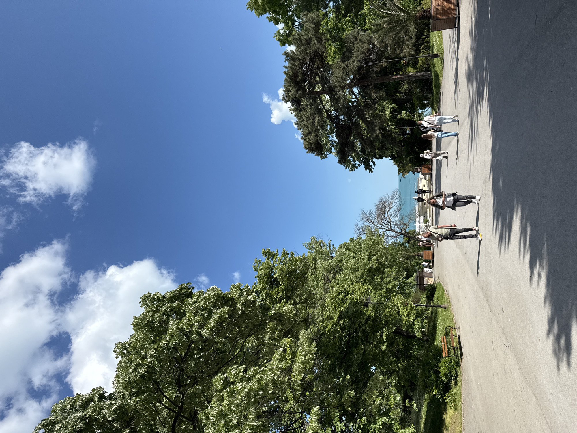 A slightly angled, sunlit shot captures a paved pathway bordered by trees and glimpses of a coastline. The sky is blue with scattered clouds. Several people are walking along the pathway. A bench is situated near the edge of the pathway.