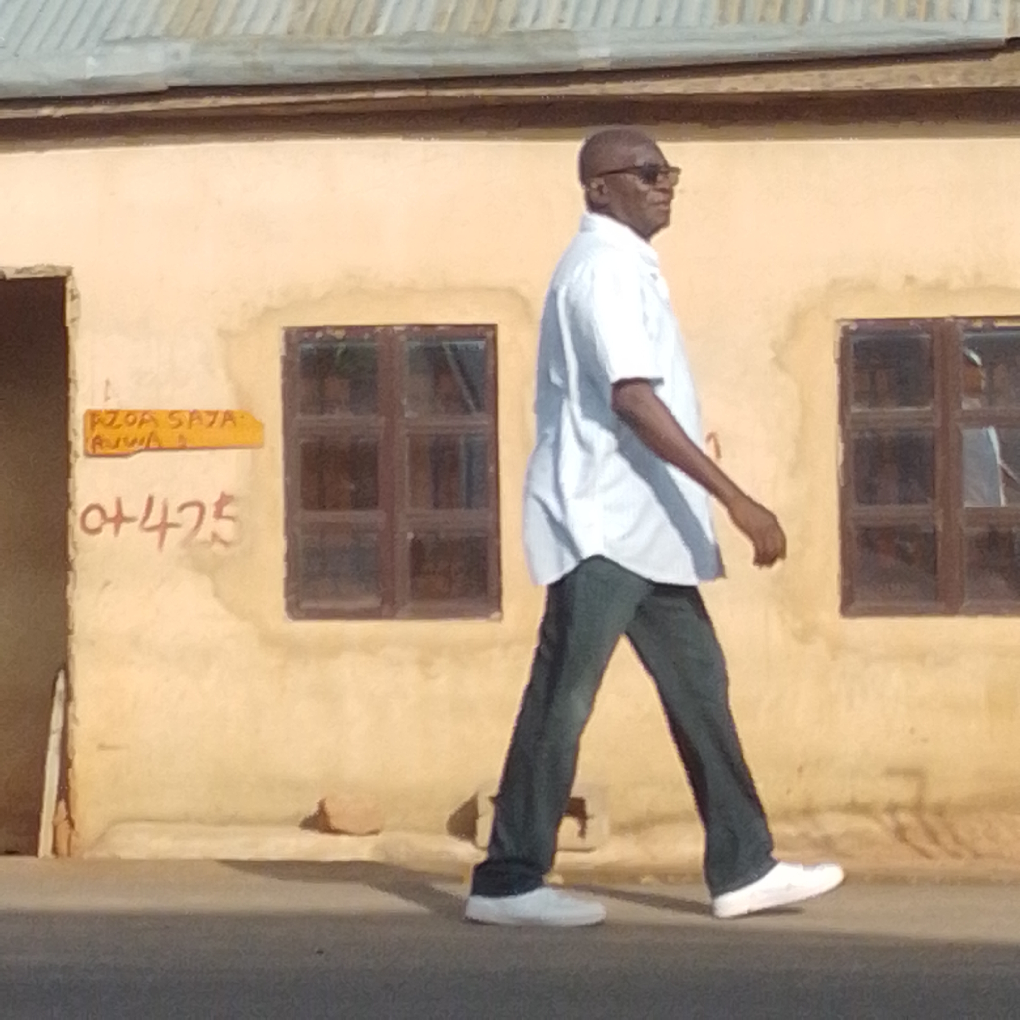 An older Black man wearing sunglasses, a white short-sleeved shirt, and dark pants walks along a paved surface in front of a pale yellow building with two dark-paned windows. A sign with orange lettering is visible on the wall to the left of the windows, and below it, the numbers "+425" are scrawled in red. The building has a corrugated metal roof. The image appears to be taken in the city of Gombe, Nigeria.
