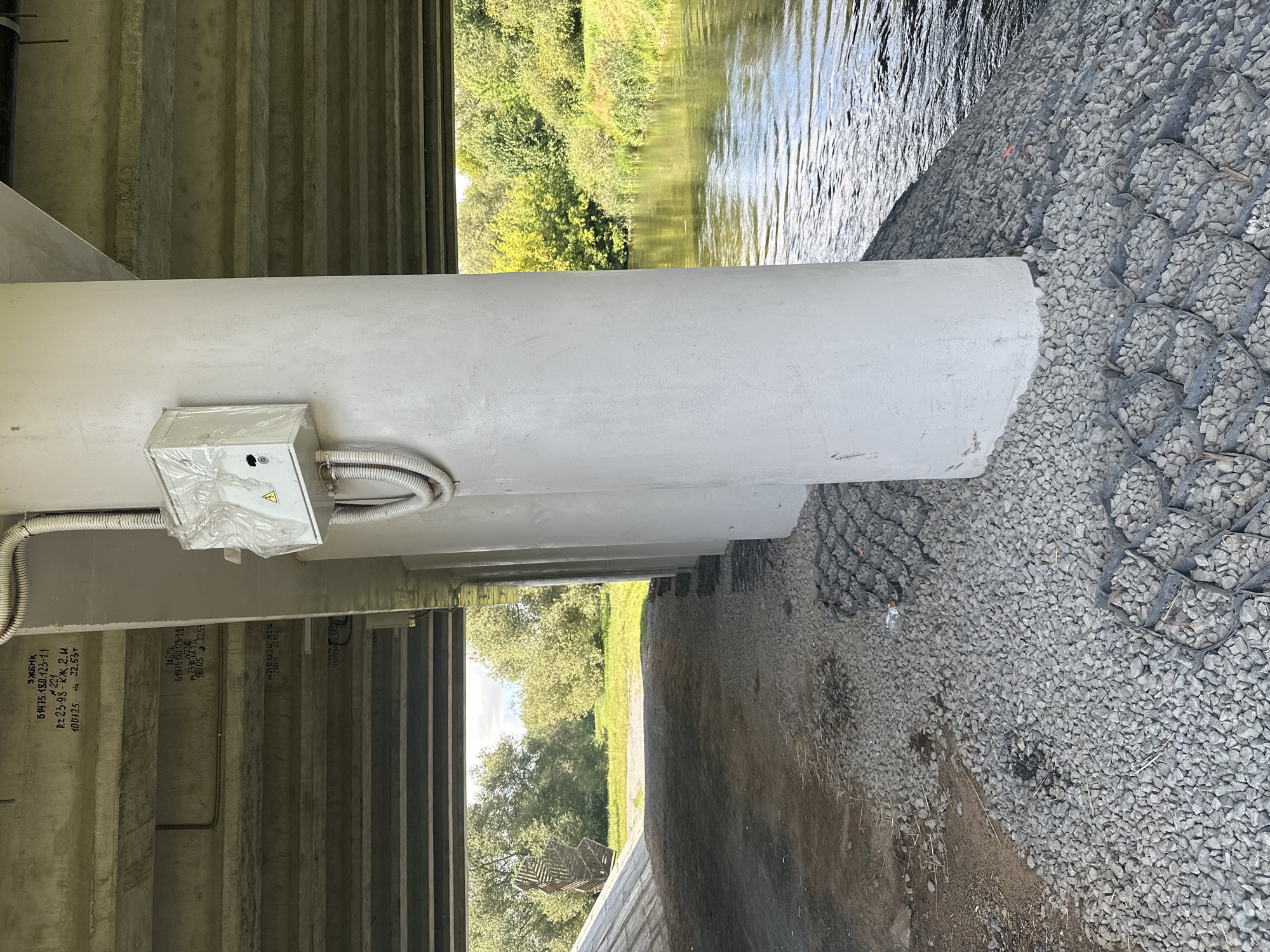 The image shows a concrete pillar supporting a bridge over a river. On the pillar, there is an electrical box covered in plastic wrap. The riverbank is covered in gravel and a grid system of some sort. In the background, there are trees and a clear sky. The text visible on the bridge's concrete surface indicates it is located in Minsk, Belarus.