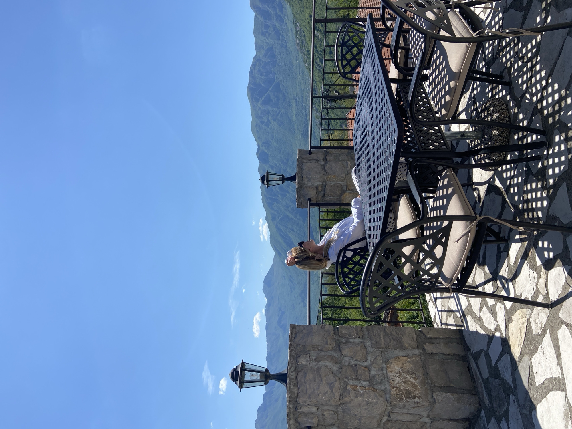 The scene is on a stone-paved terrace with a wrought-iron patio table and chairs. A woman with blonde hair is reclining in one of the chairs, her arm resting on her forehead. Behind the terrace is a stone wall and a black iron railing, beyond which lies a view of green mountains under a clear blue sky. A black lamppost stands on the stone wall. The location is Sotonići, Montenegro.