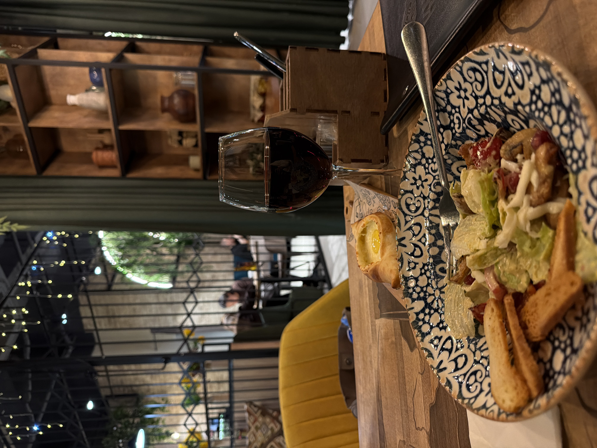 The image depicts a table setting in a restaurant, likely located in Unknown, Unknown. The table is wooden and features a blue and white patterned plate holding a salad with lettuce, croutons, and what appears to be bacon or ham. A fork rests on the plate. Next to the salad, there is a small pastry with an egg in the center. A glass of red wine sits near the plate. A wooden container holding pens or utensils is also on the table. In the background, there is a wooden shelf with decorative items and a view of other patrons seated at tables, with string lights visible above. A yellow chair is also partially visible.