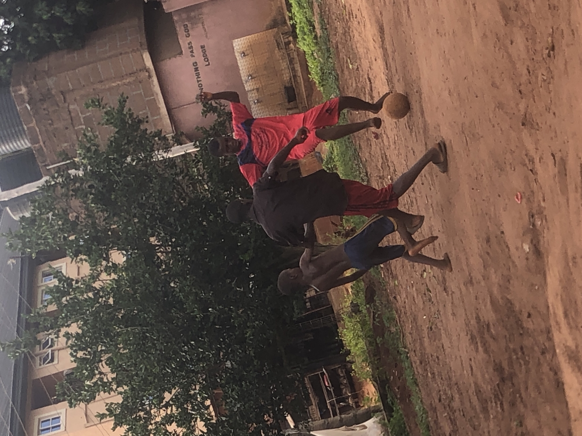 The image displays an active scene of three young males playing football on a dusty, unpaved field or street, characteristic of informal play areas in places like Ukwu-Abwa, Nigeria. The daylight setting suggests a clear day, possibly late afternoon given the soft light.
In the center, a player wearing a red and blue top with red shorts is actively engaged with a worn, brownish football. He appears to be mid-action, possibly kicking or controlling the ball. Another player, dressed in a black top and red shorts, stands slightly behind him, observing the play. Further to the left and rear, a third participant, shirtless and wearing blue shorts, is also part of the game. Some of the players are barefoot, while others wear flip-flops.
The ground is predominantly light brown dirt, with patches of green foliage, including bushes and a large tree, framing the left side of the scene. In the background, several buildings are visible. One notable light-colored building, partially obscured by greenery, prominently displays vertically oriented text: "NOTHING PASS GOD" above "LODGE." This suggests the structure might be a guesthouse or a residential building with a faith-based name. The overall atmosphere conveys a lively, everyday moment within a local community.