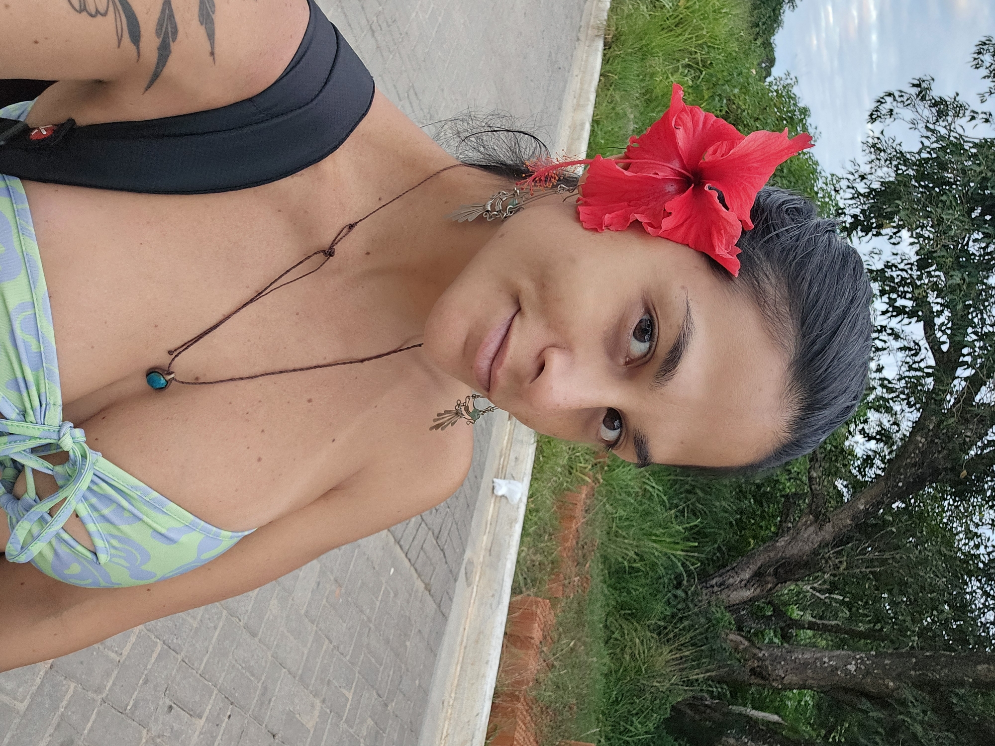A medium shot, taken at a slight angle, presents a woman in what appears to be Armação Dos Búzios, Brazil. She is wearing a blue and green floral patterned bikini top, with a black strap over her shoulder. A brown necklace with a turquoise stone pendant hangs on her chest. In her hair, she wears a vibrant red hibiscus flower, and silver earrings dangle from her ears. The background shows a paved walkway, green grass, and a tree.
