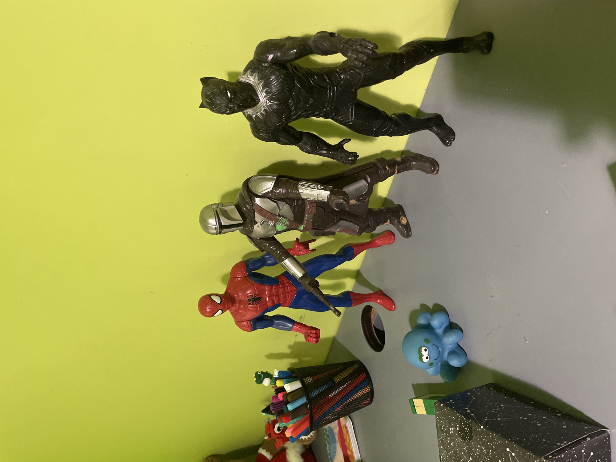 The image shows a collection of toys and objects on a surface divided into yellow and gray sections.
From top to bottom, the toys are: a Black Panther action figure, a Mandalorian action figure, and a Spiderman action figure holding a silver object. To the right of Spiderman is a small blue octopus toy. Below Spiderman is a black pencil holder filled with colorful markers and crayons. Partial views of other objects are present at the bottom, including a Santa Claus figure and a box with a constellation pattern.
The location in the city of Unknown, Unknown is indeterminate.