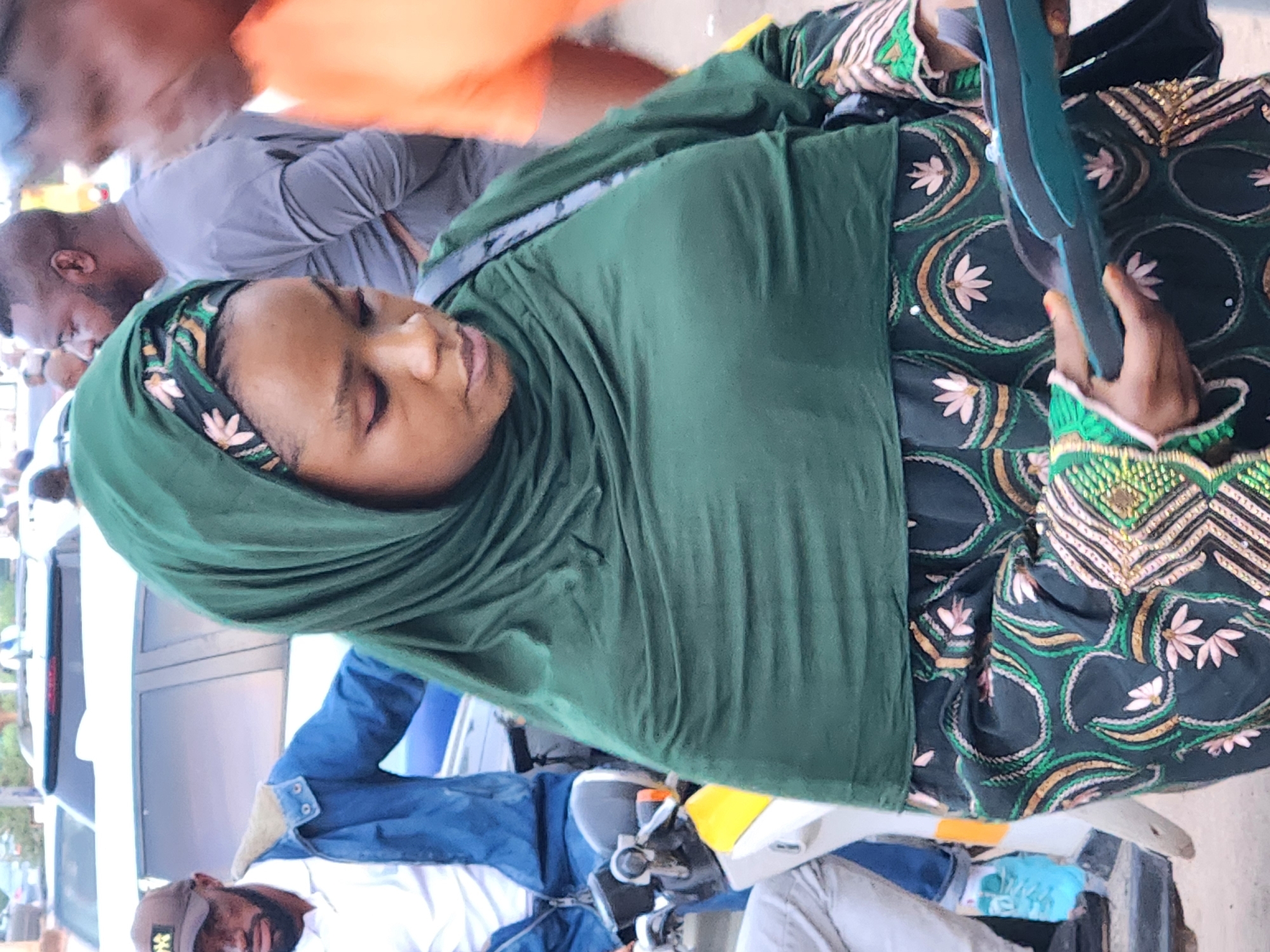 The image, oriented sideways, primarily features a woman dressed in a dark green hijab and a long-sleeved patterned dress. The dress has a dark base adorned with intricate circular and floral motifs in green, gold, and light pink or white. She holds a dark teal object, possibly a smartphone or small tablet, in her right hand. Her gaze is directed downwards, and her expression appears neutral.
Behind and above her, a man in a light grey t-shirt is partially visible, seen from the back, with a short beard. Further back, another blurred figure wears an orange shirt. Below the woman, another man wearing a white shirt and a dark baseball cap is seen seated on what appears to be a motorcycle or scooter, with blue jeans and bright blue sneakers visible. His face is also angled downwards.
The setting is an outdoor public area, likely a street or a gathering place, indicated by the presence of multiple individuals and parts of vehicles, including a white car or van in the background. The lighting is bright and natural, suggesting daytime. No discernible text is visible.