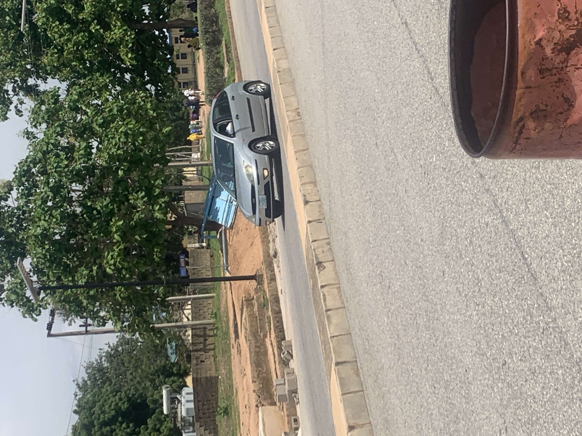 The image shows a grey sedan parked on the side of a paved road in Jalingo, Nigeria. The car is positioned diagonally to the road, with its front facing towards the left of the frame. The road itself appears to be a typical urban street, with a curb and sidewalk. To the left of the road, there is a grassy area with several trees, some of which are lush and green. In the background, beyond the trees, there are some buildings, including what appears to be a school or university campus with multiple stories. There are a few people visible in the background, some walking and others standing in small groups. The scene suggests a daytime setting, possibly late morning or early afternoon, given the lighting. The weather appears to be clear. A large, reddish-brown object, possibly a large pipe or barrel, is visible in the foreground on the right side of the image, partially obscuring the view of the road. There is some construction or earthworks visible on the left side of the road, with piles of sand and some building materials. No text is clearly discernible in the image.