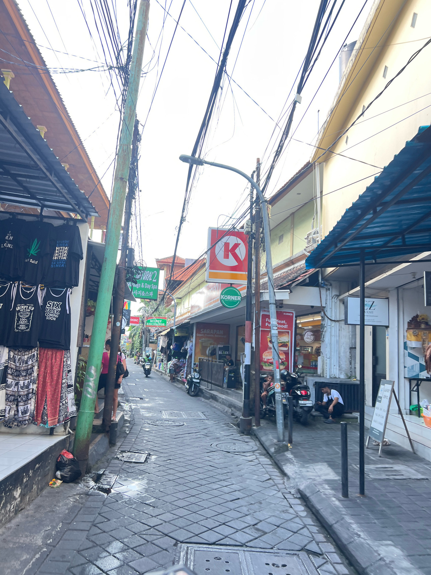 The image depicts a narrow street in Kuta, Indonesia, likely a tourist area, characterized by a cluttered overhead network of electrical wires supported by utility poles. The street is paved with dark gray square tiles and has a gutter system. Shops line both sides of the street, with awnings providing shade.
On the left side, a shop displays several black t-shirts with various graphics and text, including one with a marijuana leaf motif and another that reads "SIX PACK COMING SOON." Below the t-shirts, patterned fabrics, possibly sarongs, are also for sale. A green utility pole stands prominently in the foreground, with a black garbage bag and a small offering at its base. A woman in a pink top and dark shorts stands near the pole, looking down the street.
On the right side of the street, a convenience store with a red "K" logo and a green "KORNER" sign is visible. Further down, a "MONEY EXCHANGE" sign is displayed. Other shops show clothing and merchandise. Several motorcycles are parked along the street. A man in a white t-shirt and dark pants is seated on the curb, looking at his phone. The buildings are of varying heights and colors, with some featuring tiled roofs. The overall scene suggests a busy commercial area with a significant amount of overhead utility infrastructure.