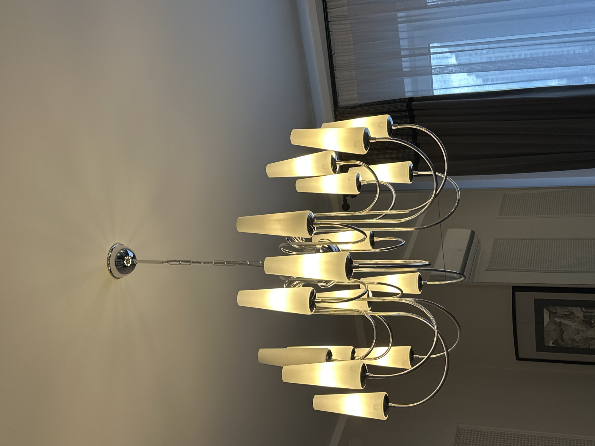This is a medium-angle, eye-level shot of a modern, multi-armed chandelier with frosted conical shades hanging from a white ceiling. The chandelier is made of polished chrome and features a central stem connected to curved arms, each holding a light fixture. The lights are illuminated, casting a warm glow on the surrounding ceiling. Below the chandelier, a portion of a room is visible, including a framed picture on a wall, a white air conditioning unit, and sheer white curtains with a dark overlay on the right side of the image, suggesting a window. The overall impression is of an indoor residential or hotel setting. The image does not provide any specific details that would allow for pinpointing its location in Minsk, Belarus.