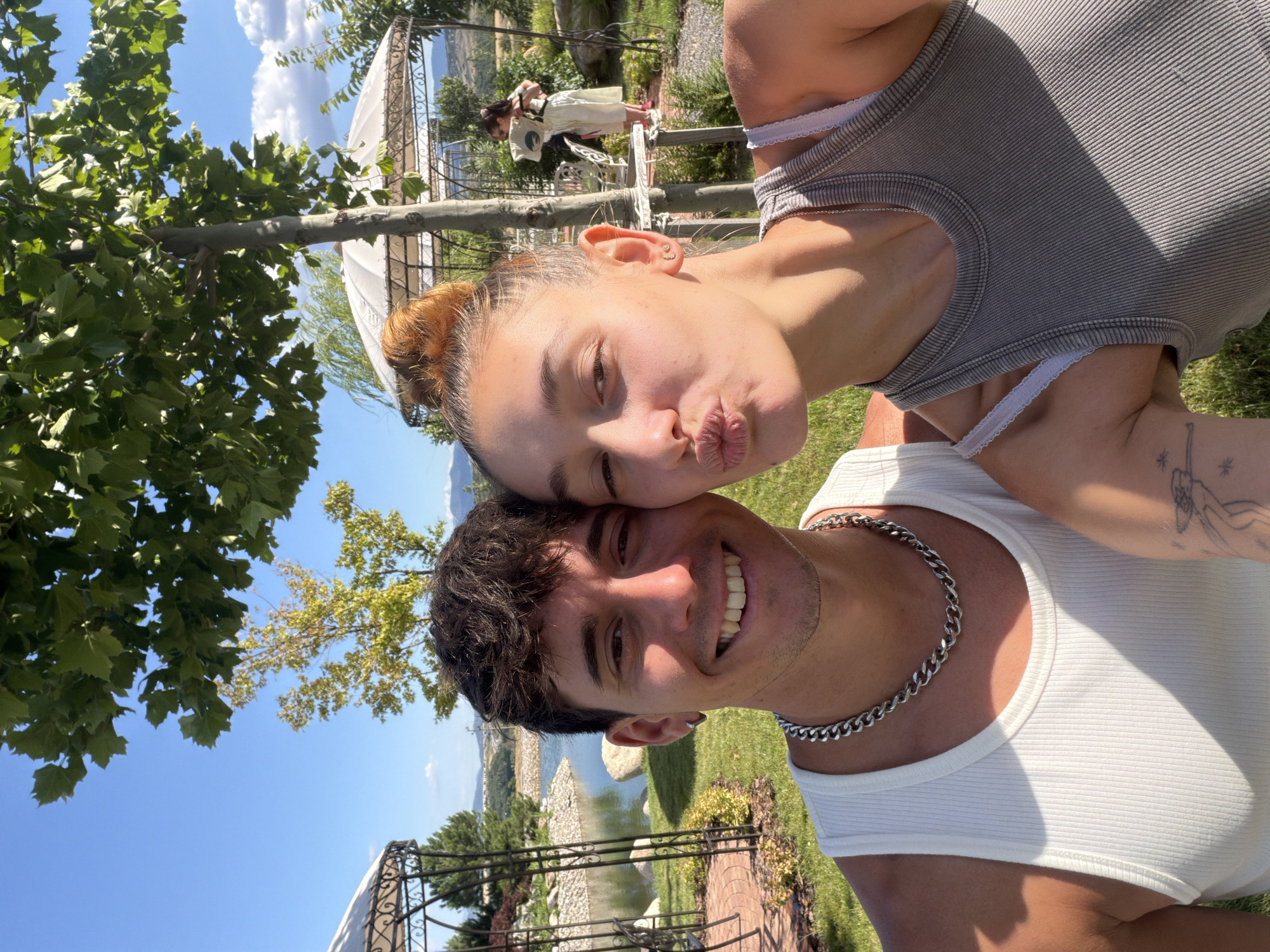 A young couple is taking a selfie outdoors. The man, on the left, is wearing a white ribbed tank top and a chunky silver chain necklace. He is smiling and looking at the camera. The woman, on the right, is puckering her lips for a kiss and looking at the camera. She is wearing a grey ribbed tank top and her hair is pulled up in a bun. In the background, there is a lake, trees, and two gazebos. Further in the background, a woman in a white dress is standing near a gazebo, holding a bag. The scene appears to be a park or a resort. The location is Kocherinovo, Bulgaria.