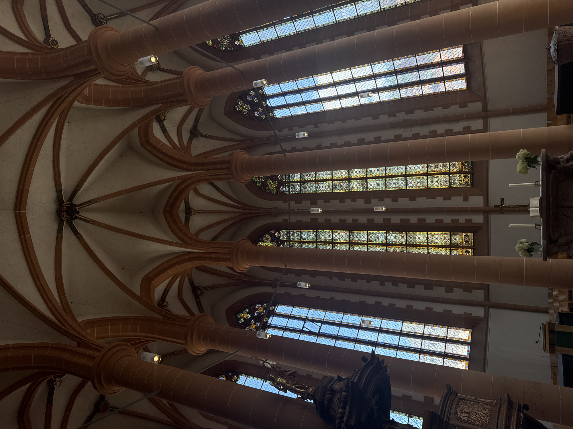 This image captures a low-angle view looking up at the vaulted ceiling and stained-glass windows of a church, likely in Heidelberg, Germany. The architectural style is Gothic, evident in the pointed arches of the ceiling and the large, multi-paned windows.
The ceiling is supported by massive, cylindrical pillars made of reddish-brown stone. Intricate stone ribs curve from the pillars, forming a complex web across the off-white ceiling. The pillars are spaced along the sides of the nave, and the windows are positioned above them, allowing natural light to filter into the space.
The stained-glass windows are a prominent feature, divided into multiple rectangular sections filled with small, diamond-shaped panes. Some of these panes are clear, showing a bright blue sky with white clouds, suggesting it is daytime with good weather. Other sections of the windows are filled with decorative stained glass, featuring geometric patterns and possibly some figural elements in muted colors of yellow, green, and blue.
Several modern light fixtures are suspended from the ceiling by wires, providing artificial illumination. One detail visible is a vase with white flowers placed on a wooden structure to the right of the frame, possibly an altar or pulpit. Another partially visible object in the lower right corner appears to be a dark, ornate wooden carving, possibly part of a choir stall or a religious statue.
The overall impression is one of grandeur and historical significance, typical of European cathedrals and churches. The interplay of natural light through the stained glass and the stone architecture creates a serene and contemplative atmosphere. There is no visible text in the image.
