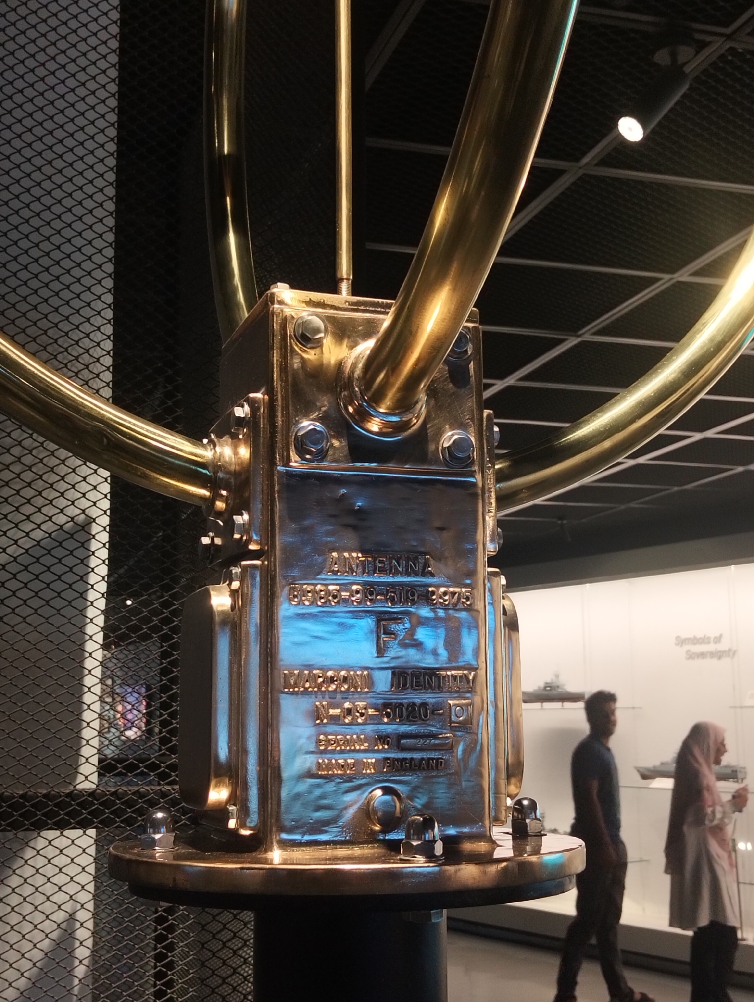 The image depicts an indoor scene within a museum or exhibition hall, likely in Dhaka, Bangladesh. The foreground is dominated by a large, highly polished brass or gold-colored metallic object, which appears to be an antique or specialized antenna. It features tubular arms extending from a central, bolted housing. Engraved text on its surface provides details: "ANTENNA", "6935-99-519-9975", "F2", "MARCONI IDENTITY N-05-5020-10", "SERIAL NO" (followed by an indistinct number), and "MADE IN ENGLAND".
In the mid-ground, two individuals are walking through the exhibition space, slightly out of focus. A man wearing a dark t-shirt and trousers is on the left, while a woman in a light-colored abaya and a pinkish-beige hijab is on the right, appearing to observe the exhibits.
The background features a white display wall showcasing models of ships or naval vessels. Text on this wall reads "Symbols of Sovereignty", suggesting a focus on national history or maritime power. A dark, mesh-patterned wall is visible to the left, and the ceiling has a grid structure with spotlights providing artificial lighting. The scene captures visitors engaging with a historical or technical display in a museum setting.
