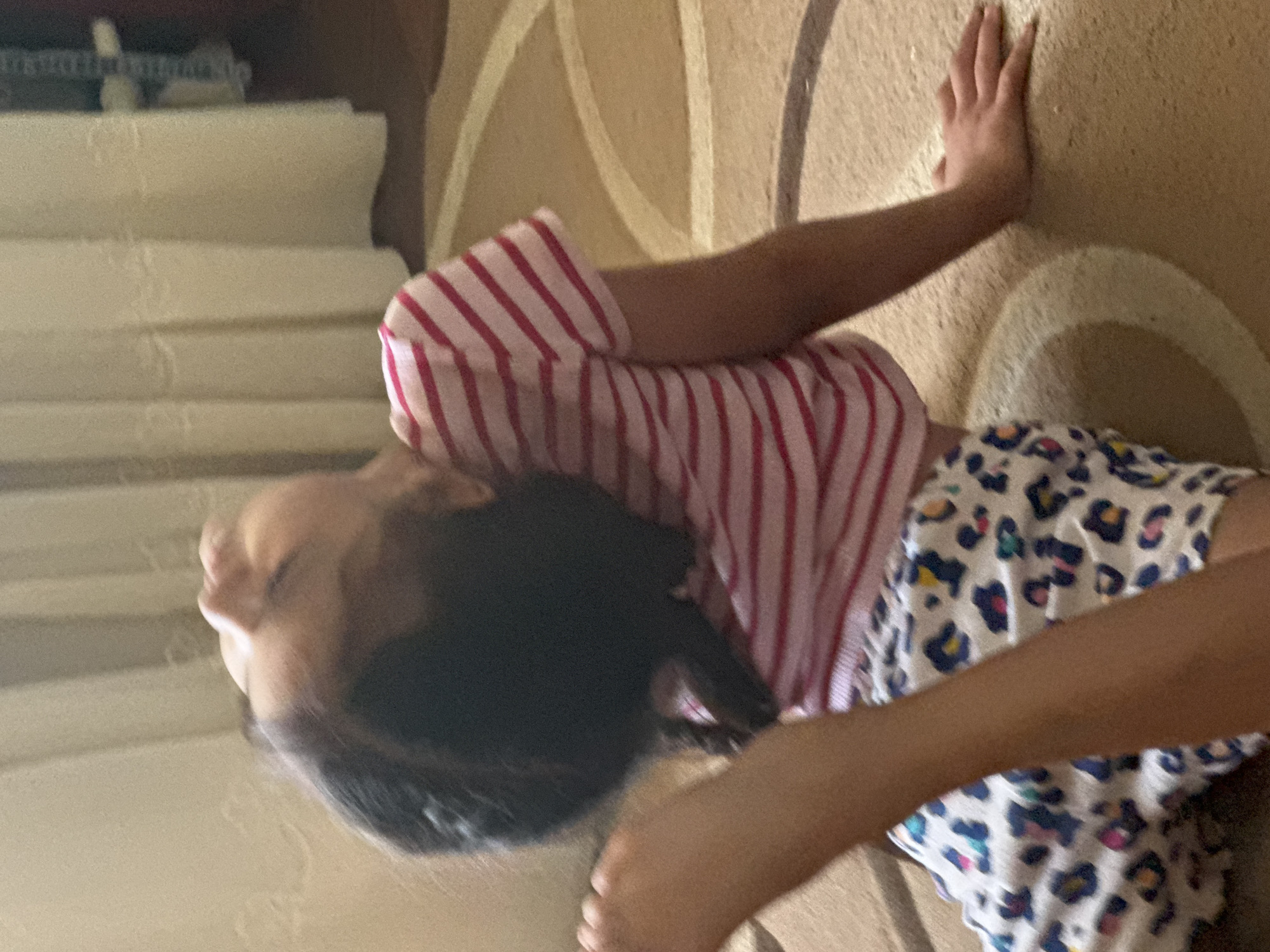 An individual, appearing to be a young girl, is depicted in an indoor residential setting within the city of Unknown, Unknown. The child is positioned on a light brown/beige carpet featuring concentric circular patterns in contrasting shades. She is engaged in a backbend or deep stretch, with her torso arched backward and her head hyperextended, orienting her face upwards. Her eyes appear closed or partially closed. She wears a short-sleeved t-shirt with horizontal red and white stripes and white shorts adorned with a multi-colored animal print (blue, pink, yellow, black spots). The child's right hand is placed flat on the carpet for support, while her left arm is partially cropped from the top-left of the frame. Her left leg is bent at the knee, with the foot raised and partially visible. In the background, light beige, vertically pleated curtains are visible, suggesting a window covering. The image exhibits a slight blur and noticeable grain, consistent with ambient or low-light photography.