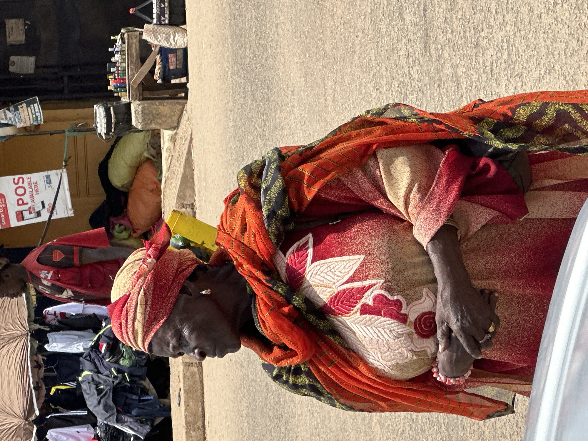 In Bukuru, Nigeria, a woman stands on a dusty street, her gaze directed left. She wears a vibrantly patterned headscarf and a flowing red and beige dress adorned with floral embroidery. A bright orange and black scarf is draped over her shoulders. Her hands are clasped in front of her, with a beaded bracelet on her left wrist and rings on her fingers. Behind her, a market stall displays various goods, including clothing, sacks of items, and bottles of products on a wooden table. A man in a red vest stands to the left of the stall. The ground is a mix of packed dirt and asphalt.