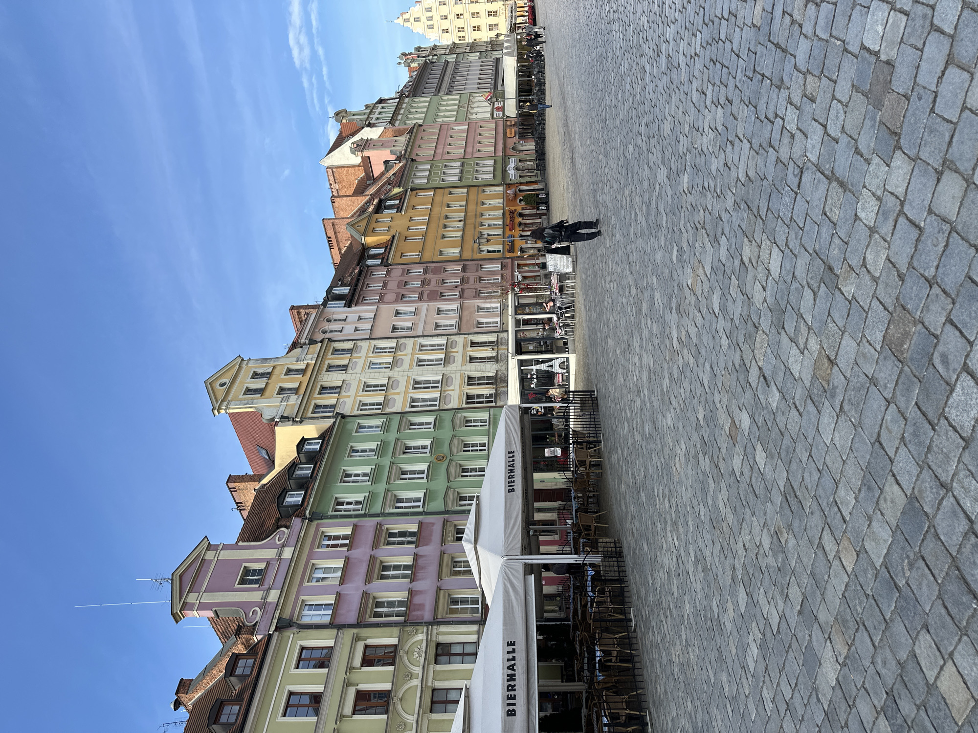 This is a street scene in Wrocław, Poland. The image shows a row of tall, colorful buildings with multiple windows and varied architectural designs lining the street. The buildings are painted in hues of yellow, pink, green, and other pastel colors. In front of the buildings, there's an outdoor seating area with tables and chairs, likely belonging to a restaurant or cafe. An awning with the word "BIERHALLE" is visible. The ground is paved with gray cobblestones. Two people are seen walking on the cobblestone street. The sky above is blue and clear.