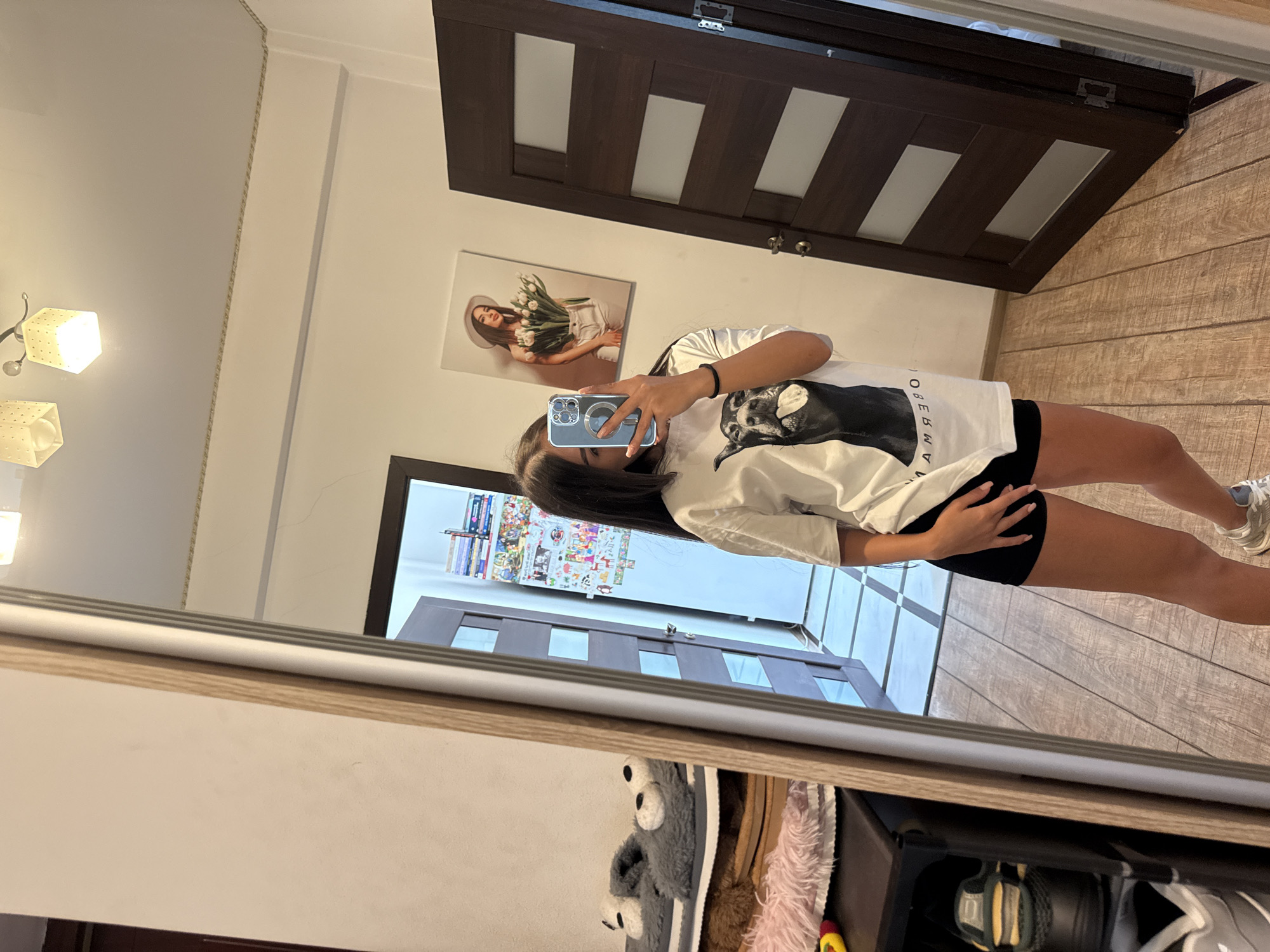 A young woman is taking a mirror selfie in what appears to be a bedroom. She is wearing a white t-shirt with a graphic print of a dog and black shorts. She is holding a smartphone in her right hand. The room has wooden floors and a closed dark wood door with frosted glass panels. A painting of a woman holding tulips hangs on the wall. To the left, a portion of a wardrobe or shelving unit is visible, holding slippers and other items. The overall impression is a casual, indoor domestic setting.