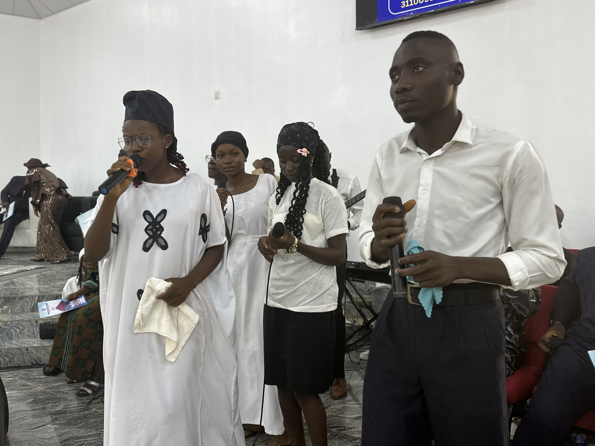 The image depicts a group of people in what appears to be an indoor event, likely a church service, in Goni Gara, Nigeria. Four individuals are standing prominently in the foreground. A woman on the left is wearing glasses, a black head covering, and a white dress with black floral embroidery. She is holding a microphone in her right hand and a white cloth in her left. To her right is another woman wearing a white dress and a black head covering. A third woman, wearing a white shirt, a black skirt, and a head covering adorned with a pink flower, is holding a microphone in her hands. On the right, a man in a white shirt and dark pants is holding a microphone in his right hand and a small blue cloth in his left. There are other individuals in the background, some seated, and some appearing to be playing musical instruments. The setting includes what appear to be steps with seated individuals on the left, and red upholstered chairs on the right. A sign is visible on the wall in the upper background.