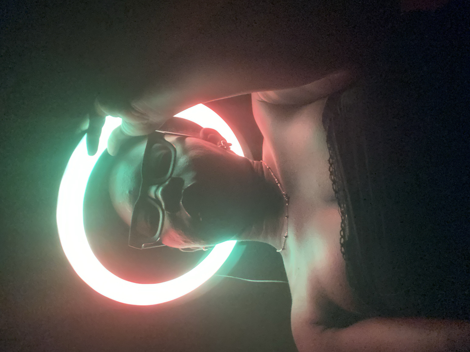 An intimate, low-light portrait features a person from the shoulders up, positioned sideways against a dark background, likely in an indoor setting. The image appears to be rotated, with the person's head oriented to the left. A prominent circular ring light, emitting vibrant green and red/orange hues, frames the person's head and neck, casting dramatic, colorful illumination.
The subject is wearing dark-framed glasses, a slender metallic necklace, and a dark garment, possibly a lacy top or bra, with its texture visible along the right side of the frame. Their skin is illuminated by the colored light, showing a range of tones. The green light predominantly shines on the left side of their face and arm, while the red/orange light highlights their collarbone, neck, and parts of their torso. One hand is partially visible at the top of the frame, near the green section of the ring light.
The person's expression is obscured by the angle and glasses, suggesting a posed shot rather than a spontaneous moment. The overall activity involves a portrait or selfie taken using a ring light for artistic lighting. The deep shadows in the background indicate night or an artificially lit environment. There are no specific visual cues to confirm the location as Maigizo, Nigeria, beyond the provided context. No text is visible in the image.