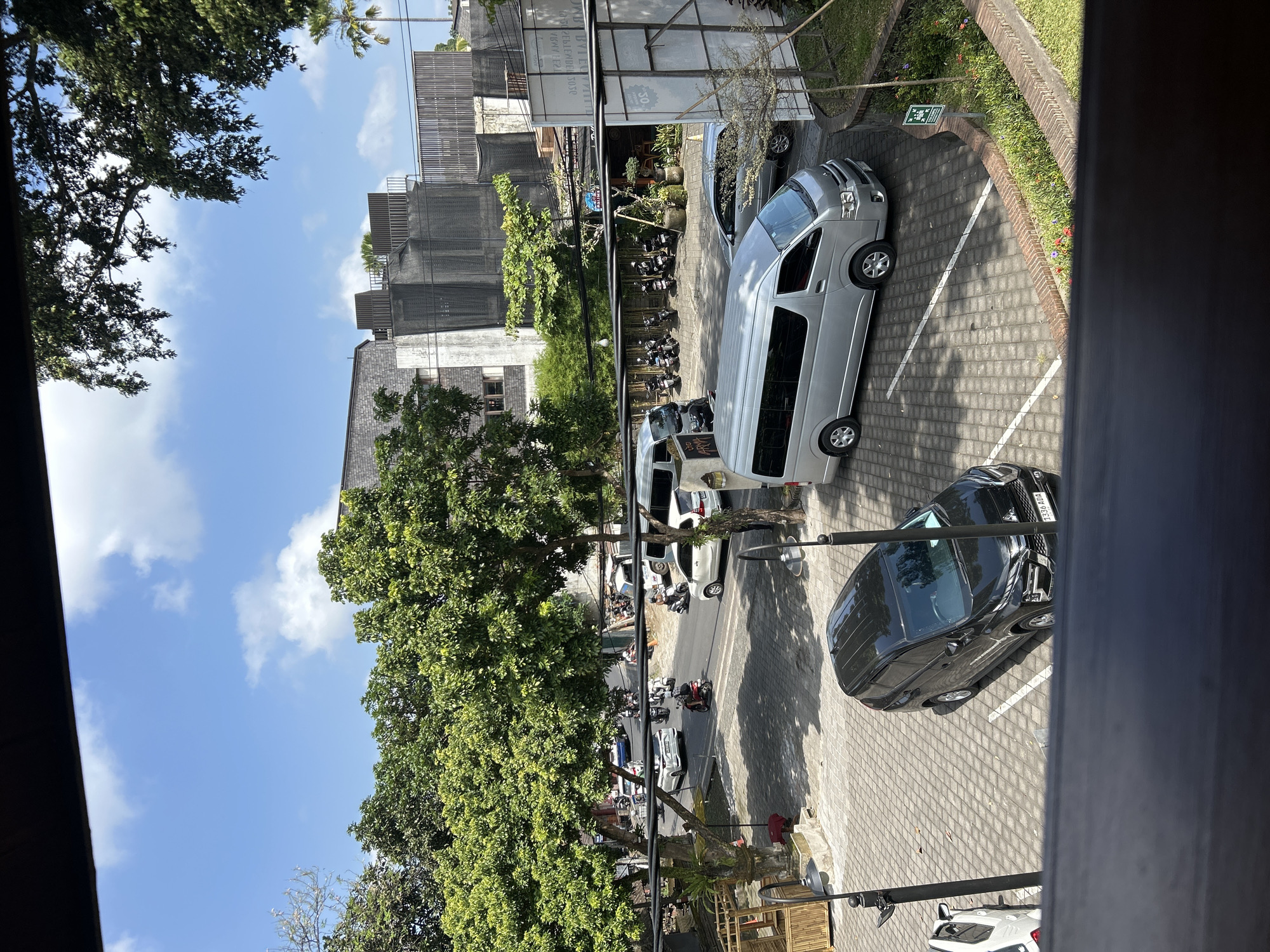 The image depicts a daytime scene in a paved parking area with a street running alongside it, likely in the city of Ubud District, Indonesia. The sky is blue with scattered white clouds. Several vehicles are visible, including a black SUV parked in a designated spot, a silver minivan parked in another spot, and multiple motorcycles and cars on the street. Trees with lush green foliage line the street and frame the buildings in the background. The buildings appear to be a mix of modern and traditional architecture, with some featuring textured gray facades and others with more open, possibly commercial spaces. A wooden structure with bamboo elements is also present on the left side of the parking area. A small patch of garden with flowering plants is situated to the right of the parking lot. Traffic, primarily consisting of motorcycles and cars, is present on the street, indicating a moderately busy urban environment. The overall impression is that of a functional outdoor space within a tropical city.