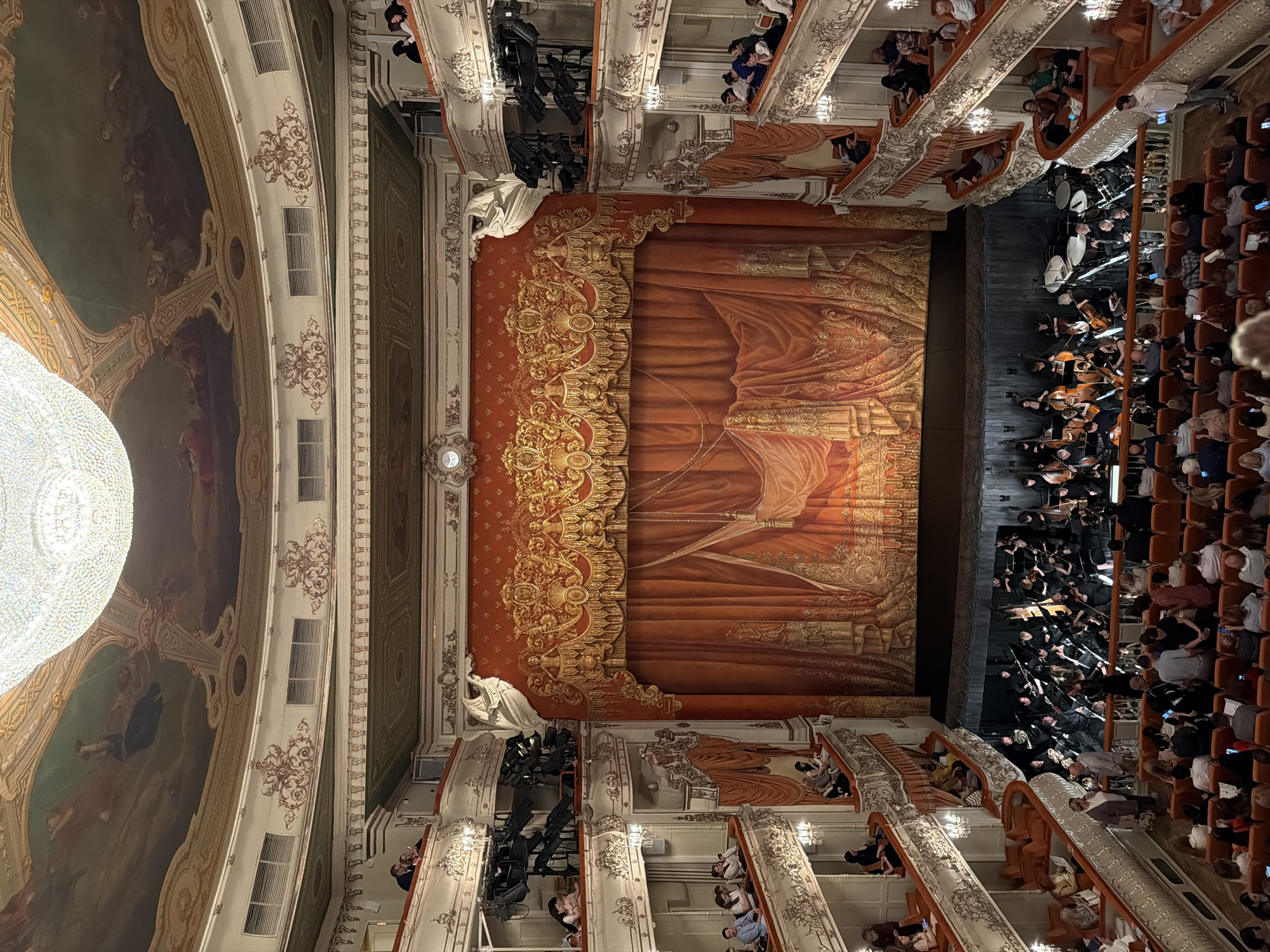 The image shows the interior of a grand theater, likely an opera house, filled with an audience and an orchestra on stage. The architecture is ornate, with multiple levels of balconies and a decorative ceiling with painted murals. A large chandelier hangs from the ceiling. The stage is set with a red and gold curtain, and an orchestra is positioned in front of it, with musicians playing various instruments, including string instruments, brass, and percussion. The audience members are seated in plush orange chairs, some are looking at their phones, while others are engaged with the performance. The overall scene suggests a cultural event taking place in a richly decorated performance space. The location is identified as the city of Lednevo, Russia.