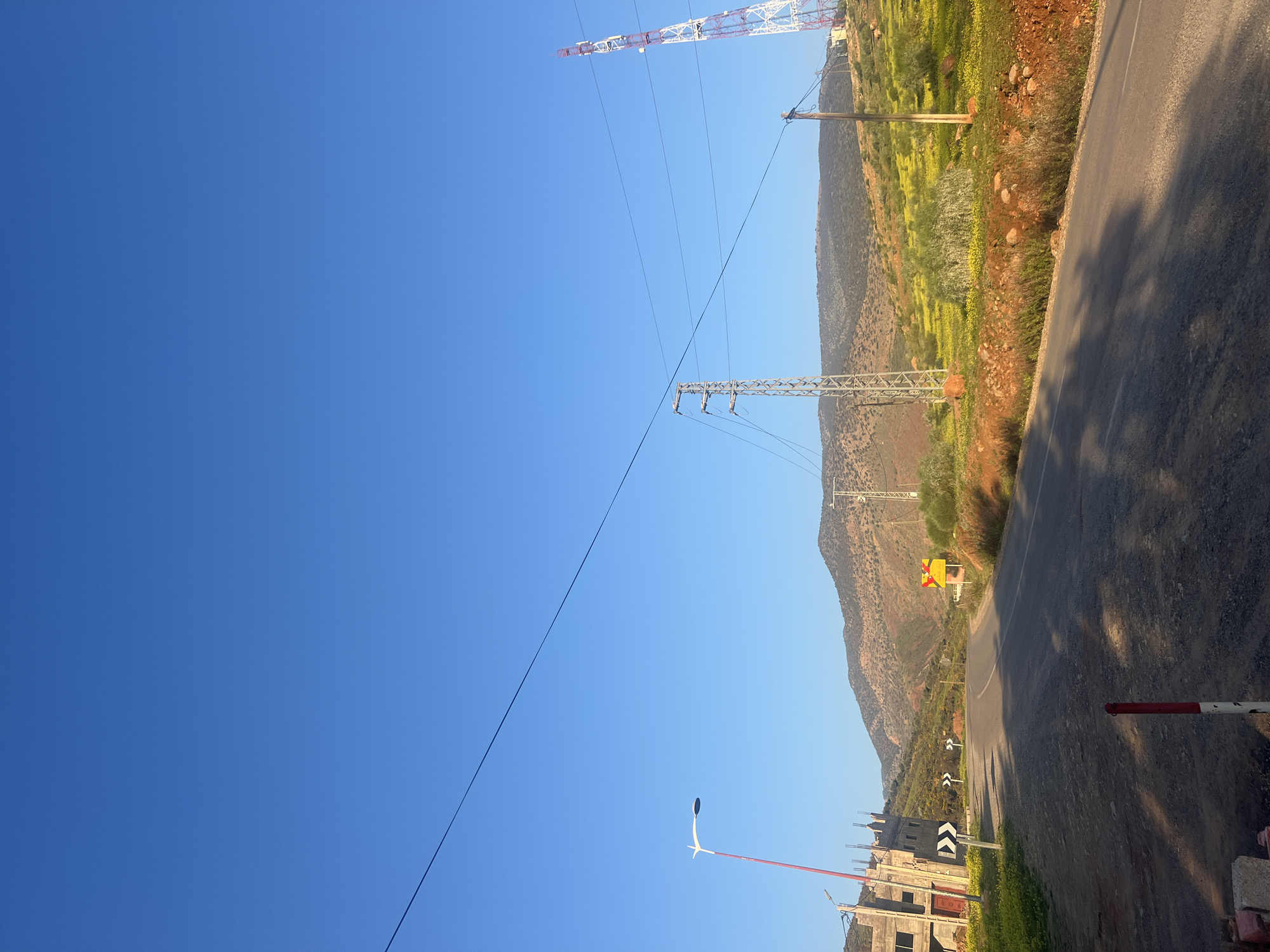The image depicts a roadside scene in Ouaouizert, Morocco, under a clear blue sky. A paved road runs diagonally across the frame, bordered by vegetation and rocky terrain. Power lines and towers are visible, stretching across the landscape. Buildings under construction and road signs are present near the road. The scene suggests a rural or semi-rural environment.
