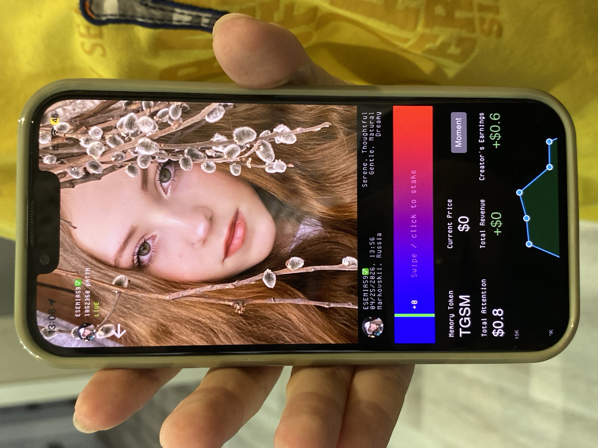 A person is holding a smartphone displaying a livestream of a young woman with hazel eyes, light skin, and light-brown hair adorned with pussy willow branches. The livestream includes the username "ESENIAS9", the date "04/25/2026", the time "13:56", and the location "Markovskii, Russia". Descriptive text includes "Serene, Thoughtful, Gentle, Natural, Dreamy." The app interface shows a Memory Token (TGSM), a total attention of $0.8, a current price of $0, and creator's earnings of +$0.6. A graph displays the creator's earnings, and the battery level of the smartphone is at 45%.