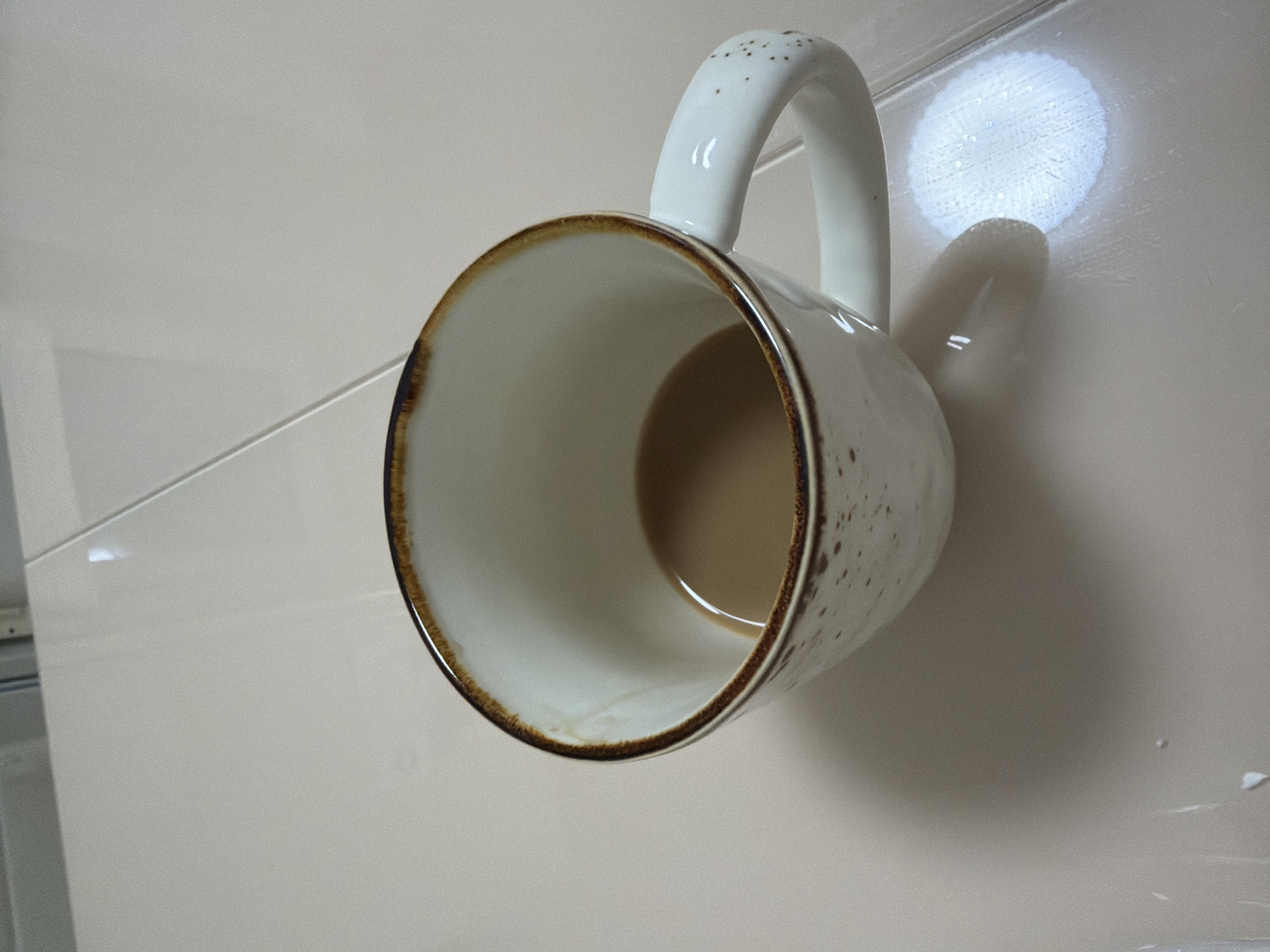 A close-up, high-angle shot shows a partially filled coffee mug with a brown rim and speckled exterior. The mug rests on a light-colored, tiled surface. The coffee inside the mug is a light brown color, and there is a subtle sheen on the liquid's surface. The background consists of the same tiled surface, with a subtle reflection of light from the mug to the right of the object. There is no indication of the specific location within Izhevsk, Russia, or any interaction with people.