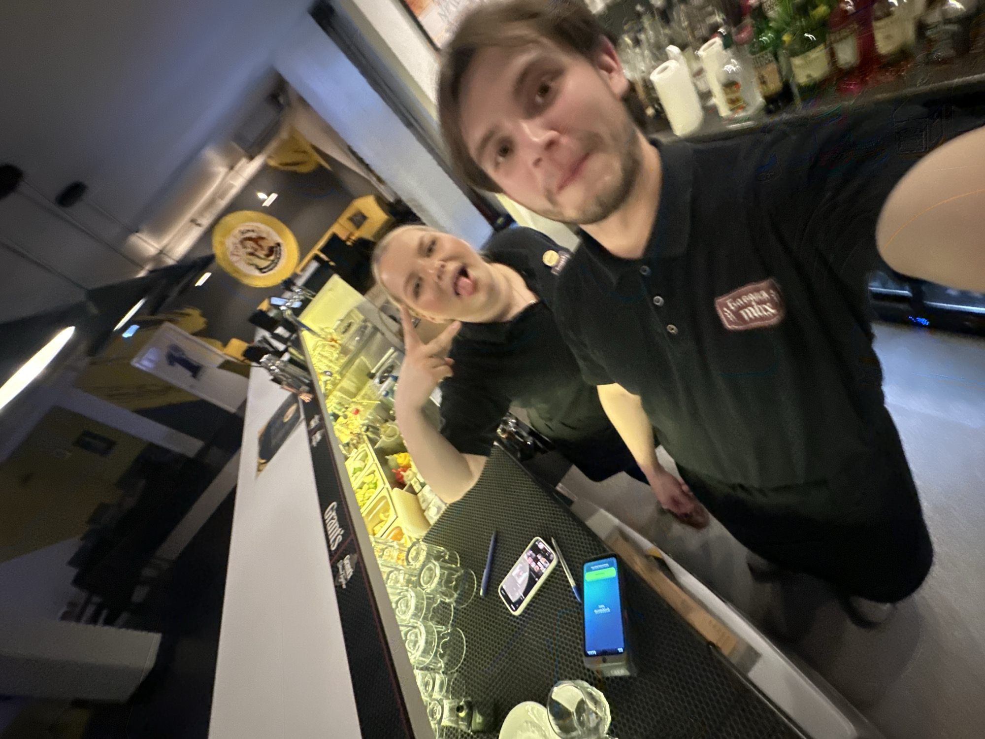 **Scene:** A slightly tilted, angled shot within what appears to be a bar setting, likely in Chaikovsky, Russia, based on the "Garage Mix" logo on the man's shirt.
**People:**
* Two individuals are present. The primary individual, a man, holds the camera for a selfie. He has light brown hair and a beard. He is wearing a dark-colored polo shirt with the "Garage Mix" logo. The other person, a fair-skinned woman with blonde hair, stands slightly behind him. She's sticking her tongue out and making a peace sign. She also wears a dark-colored shirt.
**Objects:**
* Behind the two people is a counter or bar area. There are various bottles of alcohol stacked on a shelf.
* On the counter, there is a black pad with several glasses, two phones, and two pens. One of the phones has a bright blue screen.
**Environment:**
* The location seems to be a bar or a restaurant, indicated by the bar setup and the overhead yellow lighting.
* There's a logo visible on the upper part of the wall.
* The scene is brightly lit.
**Actions/Interactions:**
* The man is taking a selfie with the woman. The woman is posing for the photo.
* The setting suggests the two people work in the restaurant or bar.